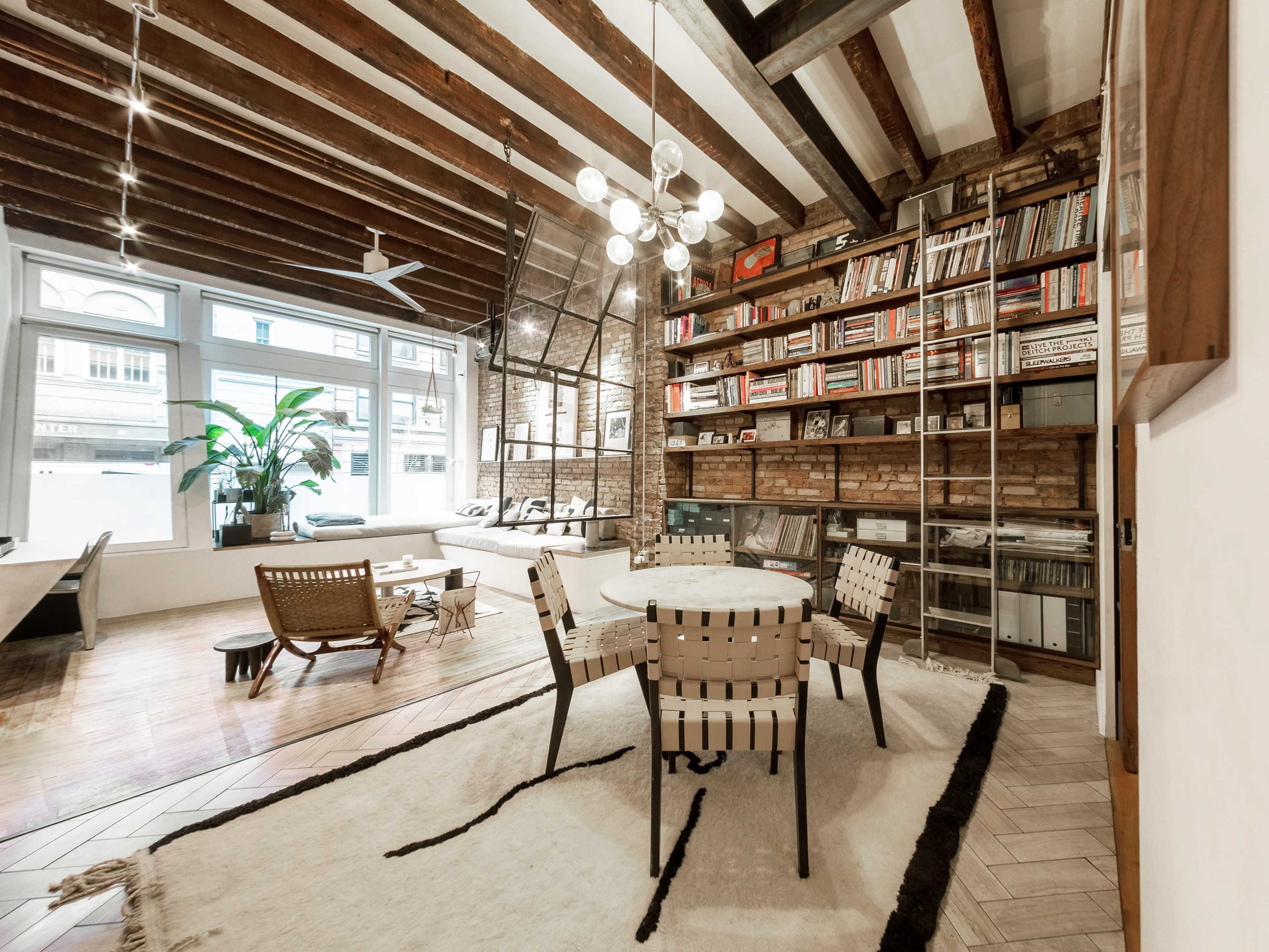 A modern living space features a circular table surrounded by four chairs, with a large bookshelf and a window showcasing natural light.