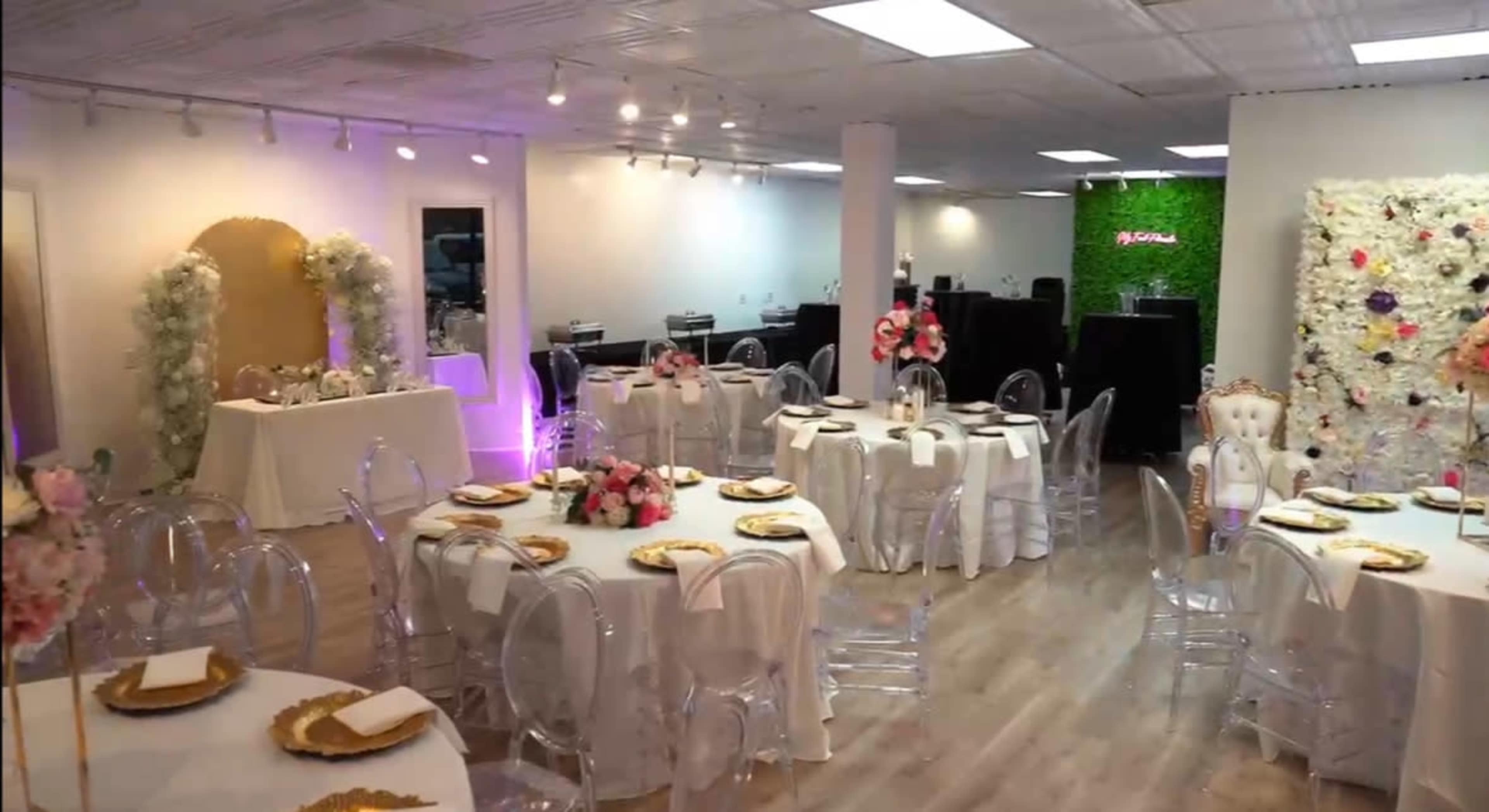 The room features elegantly set dining tables with clear chairs, floral centerpieces, and a backdrop adorned with decorations for a special event.