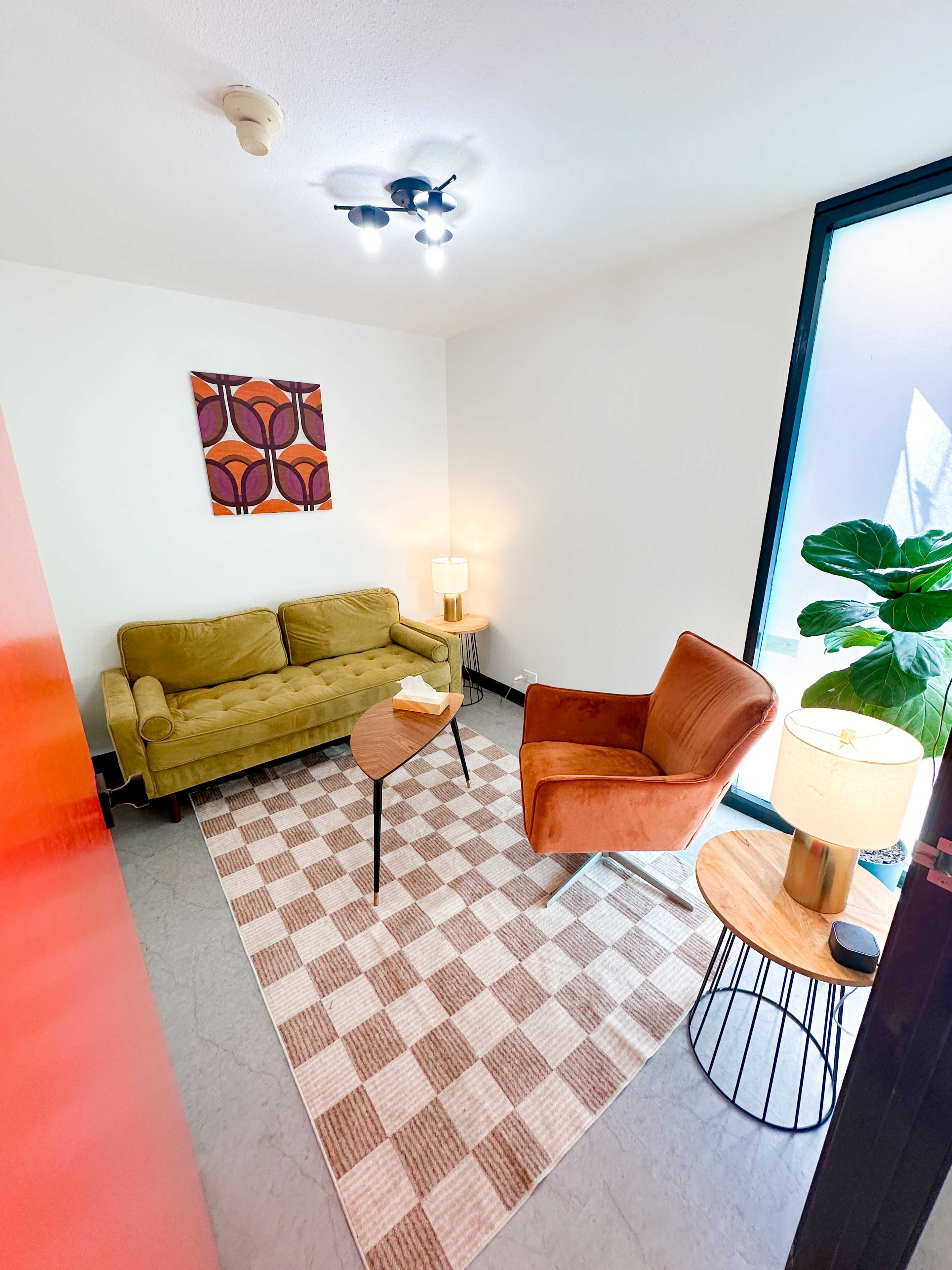 The image shows a small living space featuring a green sofa, a brown armchair, a round table, and a patterned rug.