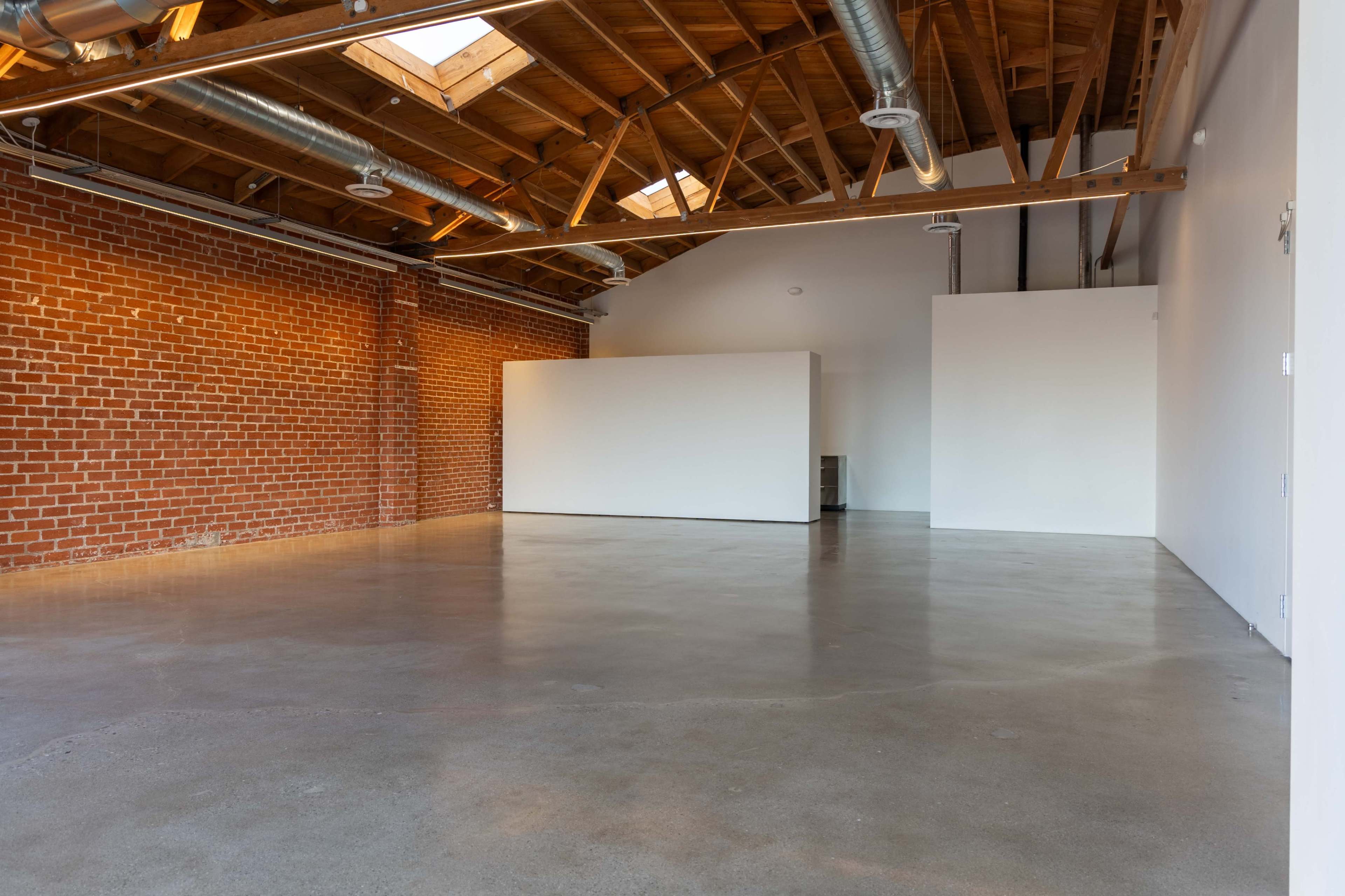 The image shows an empty industrial-style interior with exposed brick walls, a high wooden ceiling, and polished concrete flooring.