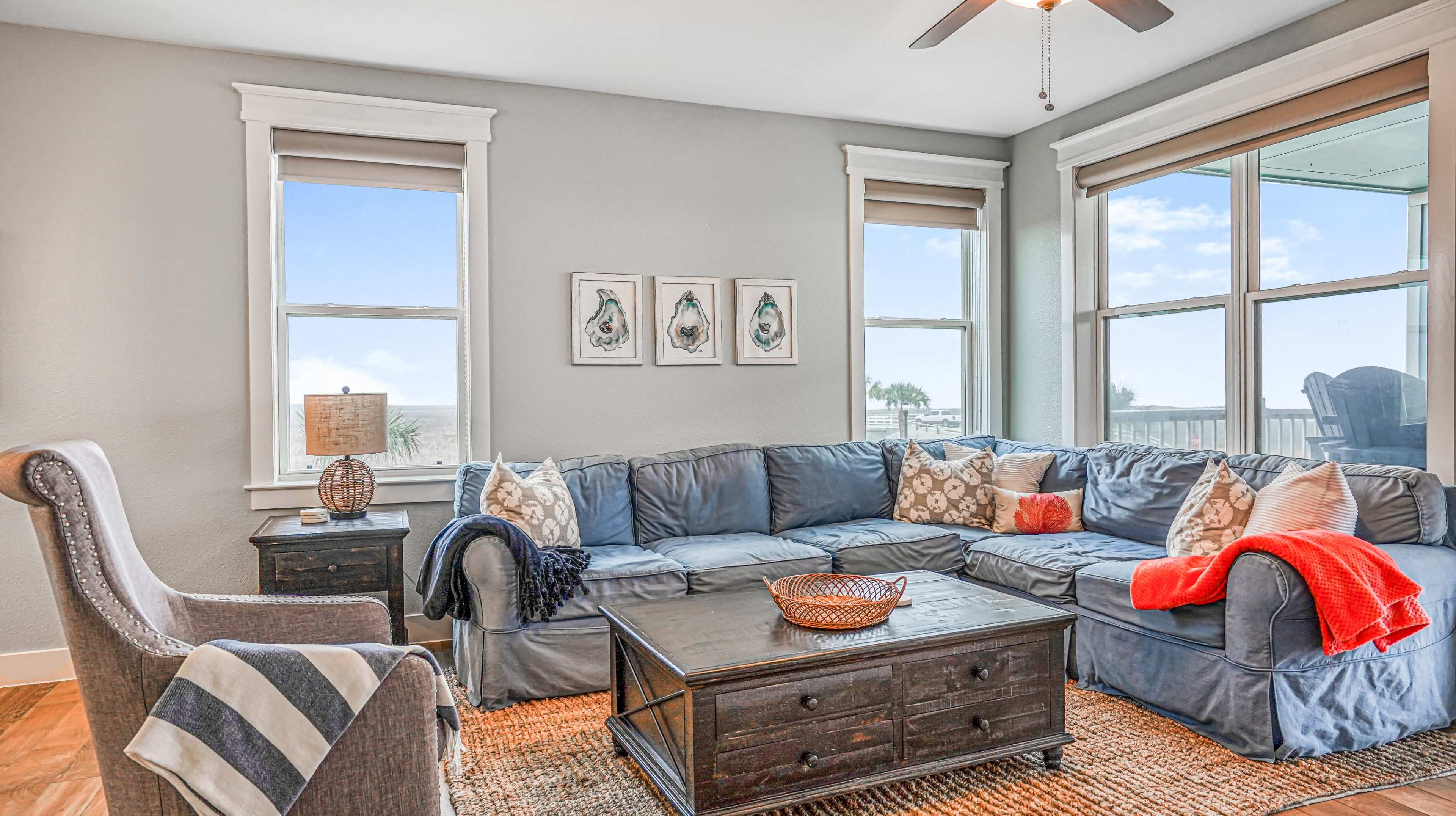 A cozy living room features a large sectional sofa with decorative pillows, a wooden coffee table, and windows that provide natural light.