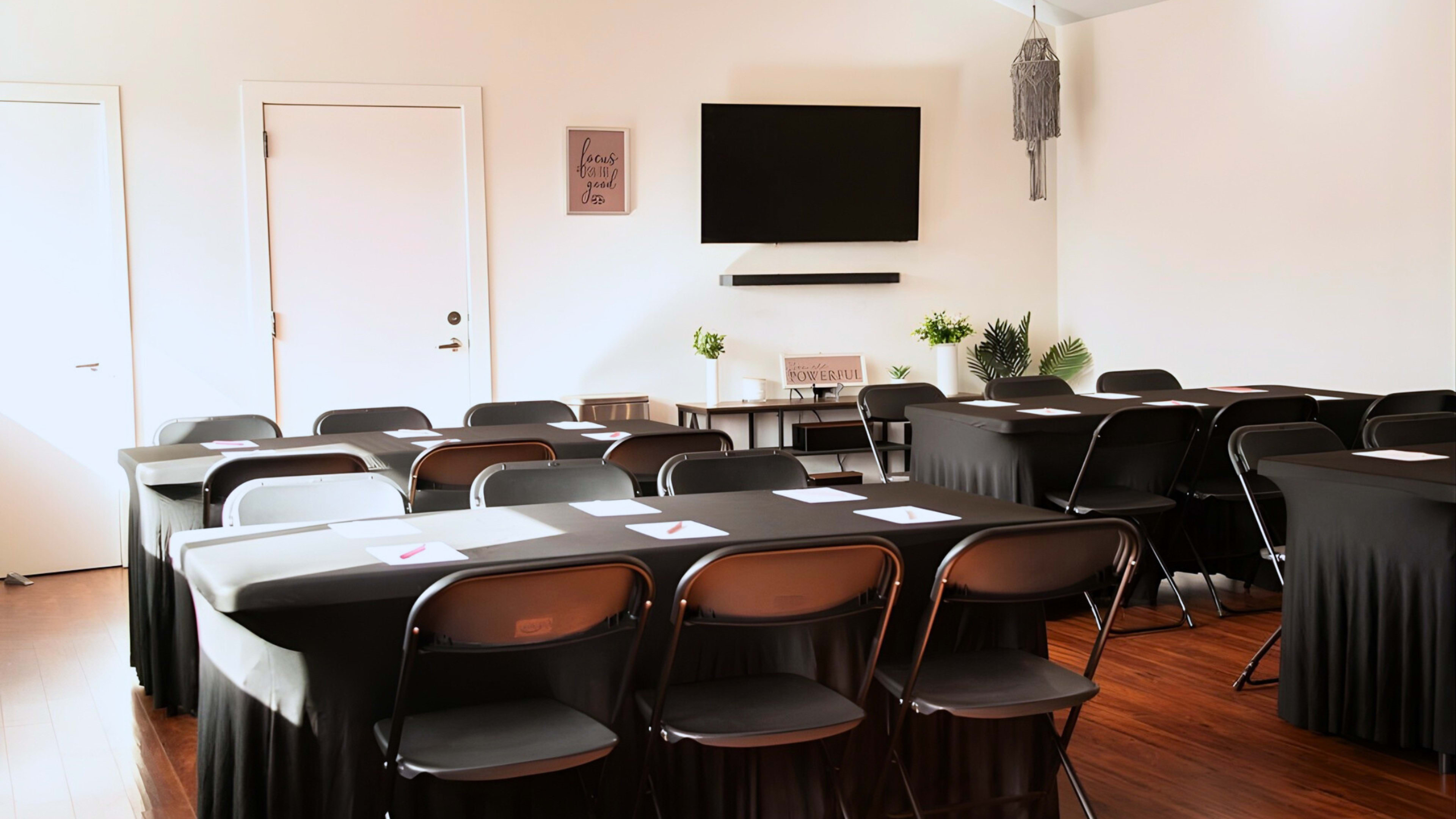 The image shows a well-organized meeting room with rows of black tablecloth-covered tables and chairs, a wall-mounted TV, and decorative plants.