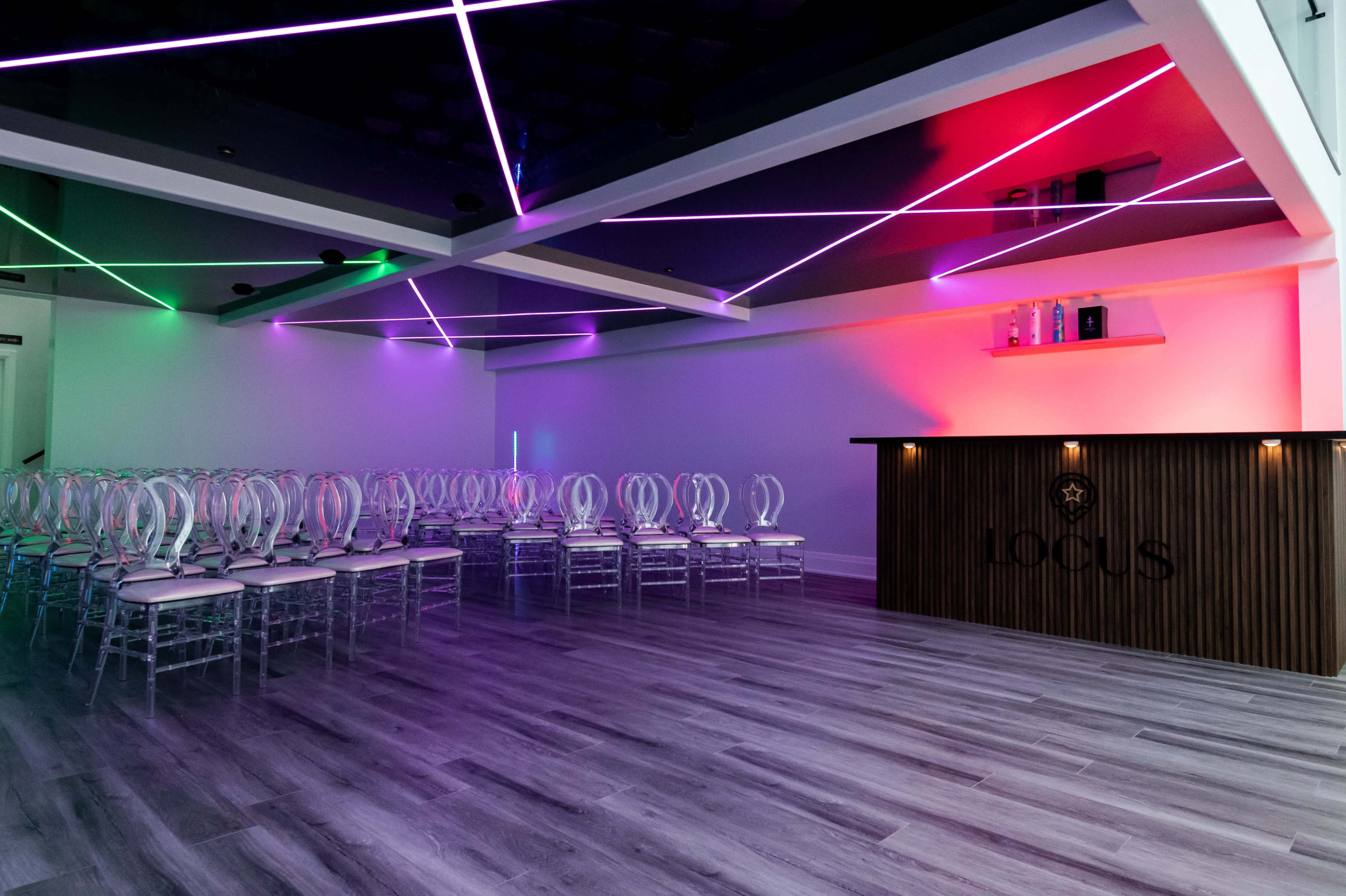 A modern event space features rows of clear chairs, a sleek wooden bar with a logo, and colorful lights illuminating the ceiling beams.