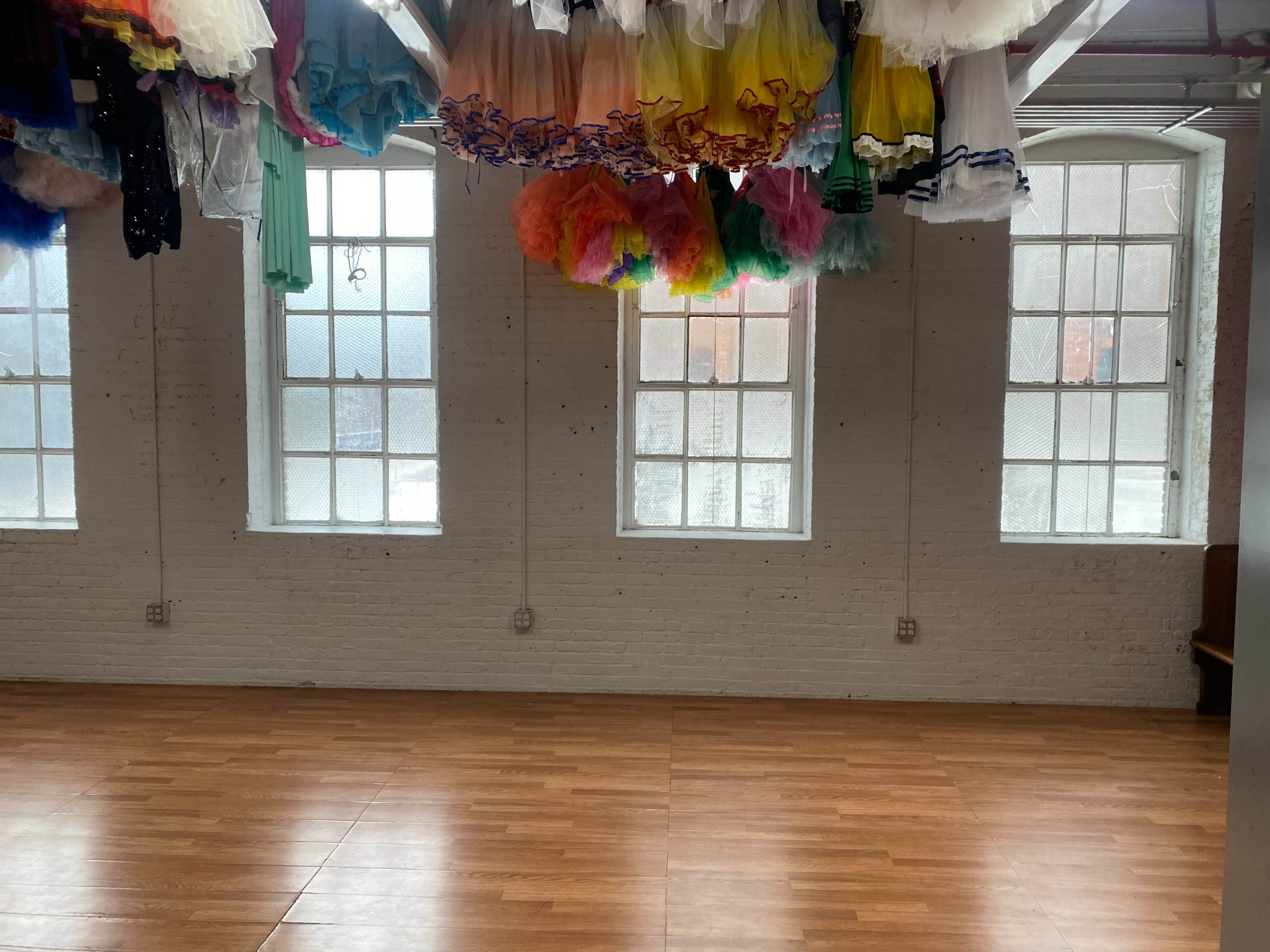 The room features wooden flooring and large windows, with colorful tutus hanging from the ceiling.