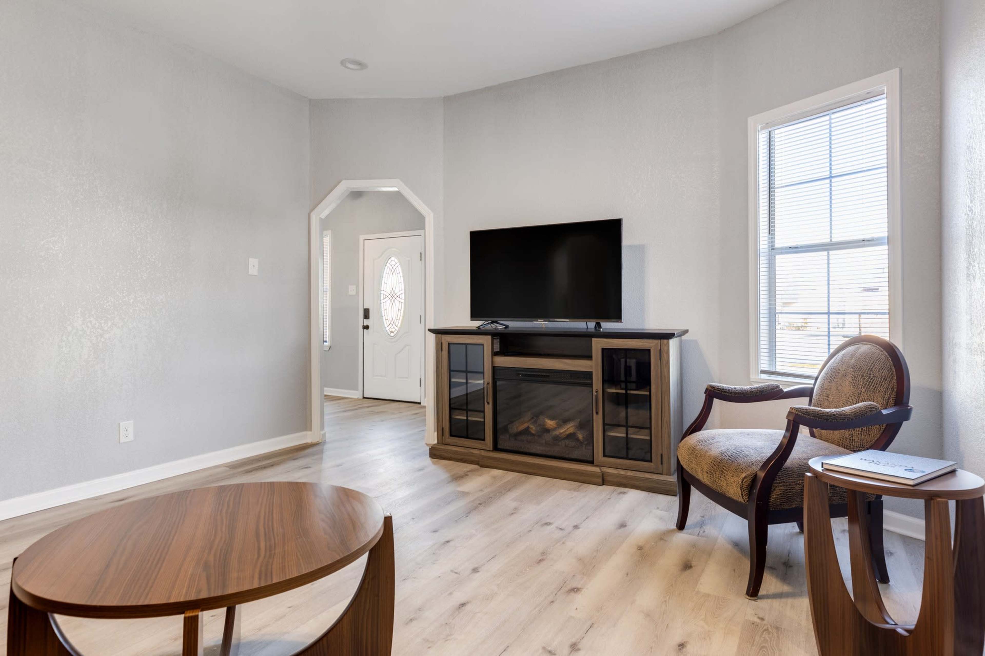 A brightly lit living room features a television on a cabinet, a coffee table, and a chair, with a door leading to another room.