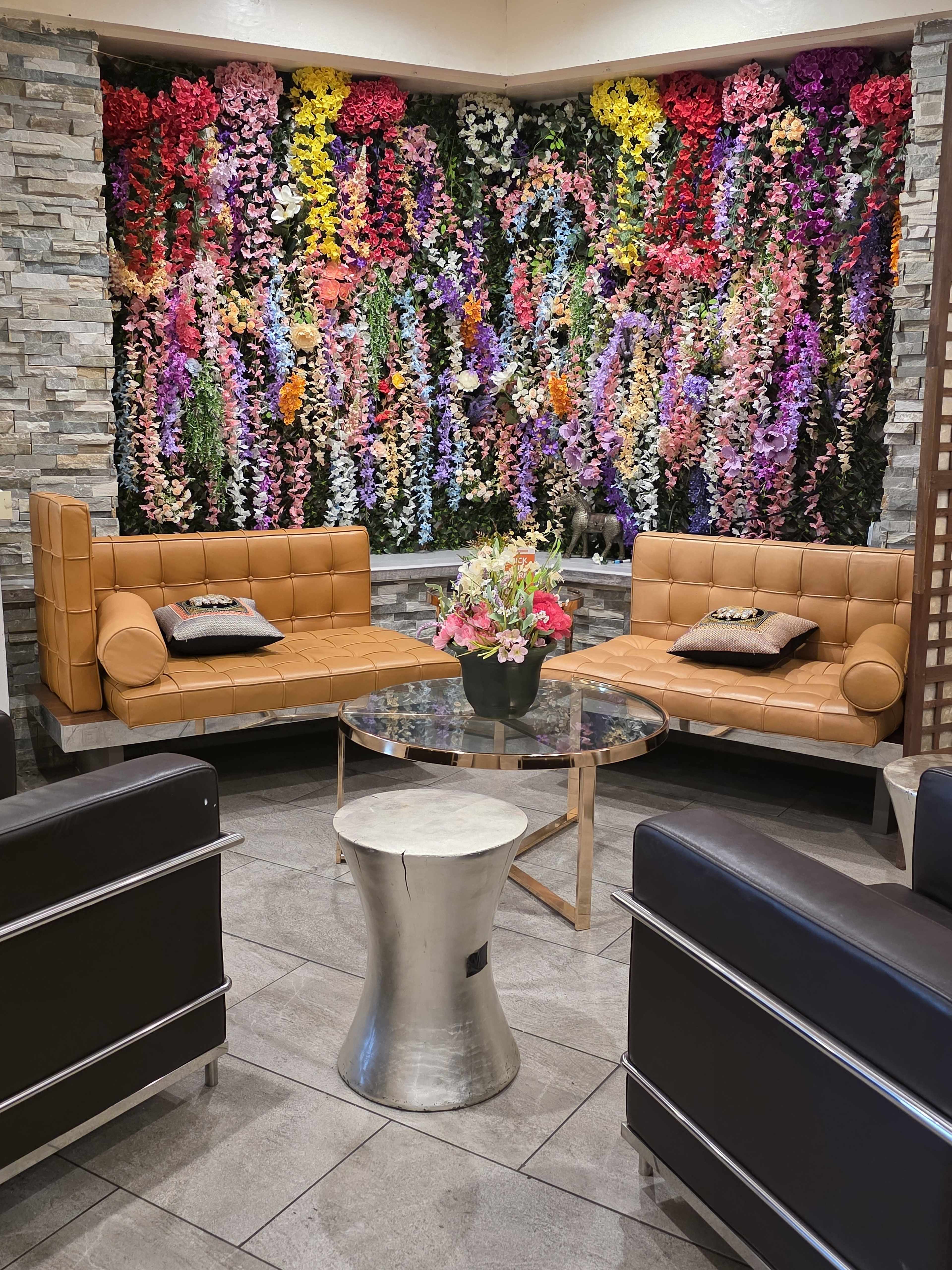 The image shows a modern lounge area with two brown leather sofas and a glass table, surrounded by a vibrant floral wall made of artificial flowers in various colors.