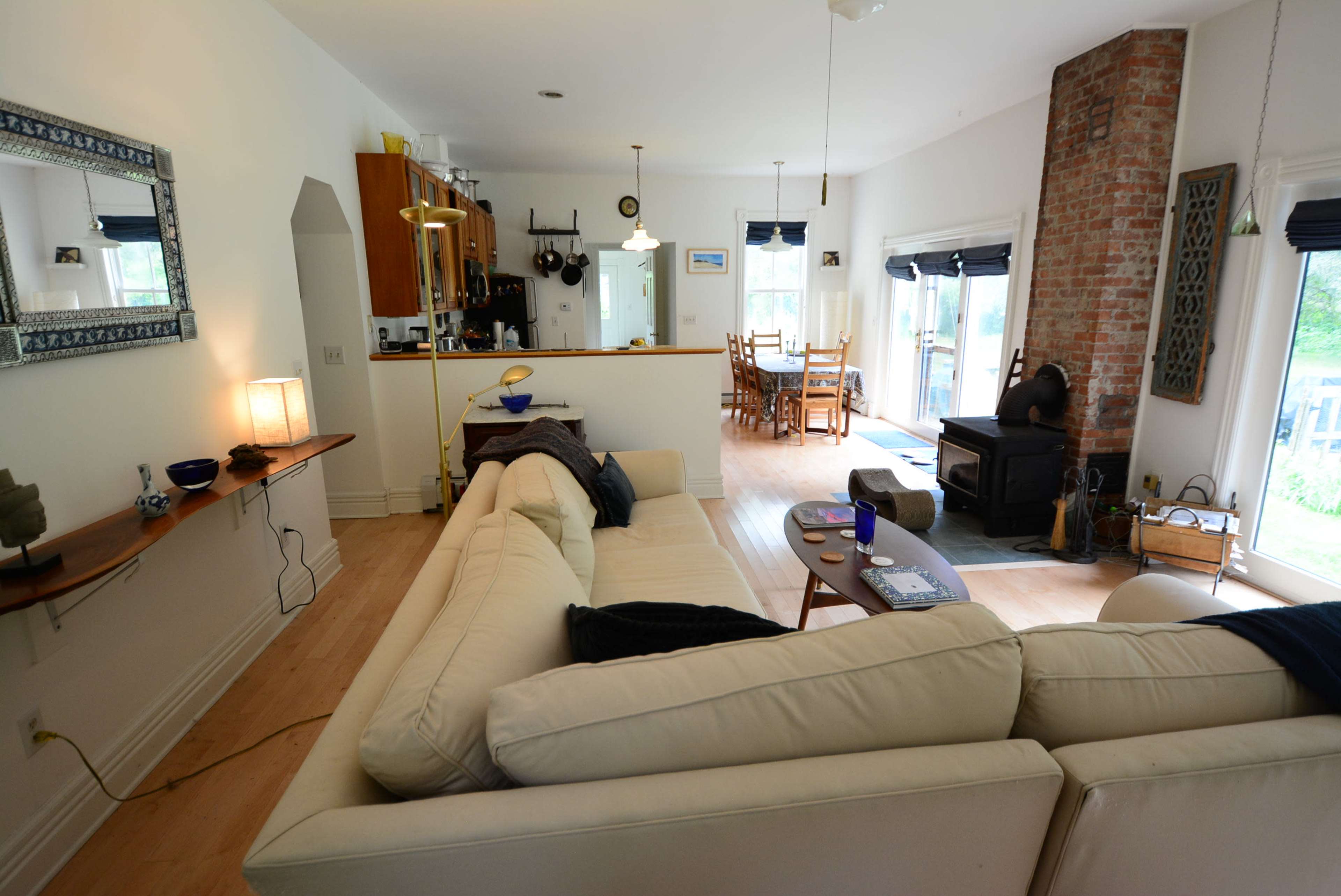 The image shows a spacious living area with a white sectional sofa facing a brick fireplace, leading into a dining space with wooden furniture and large windows.