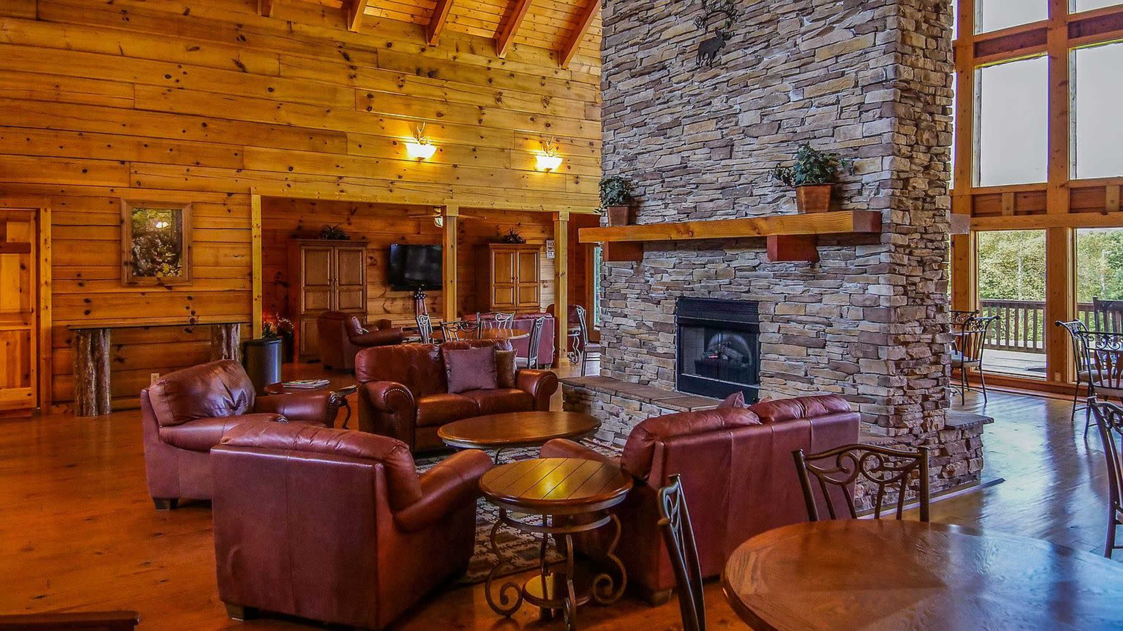 The image shows a spacious wooden interior of a cabin featuring a large stone fireplace, leather armchairs, and a dining area with wooden tables.