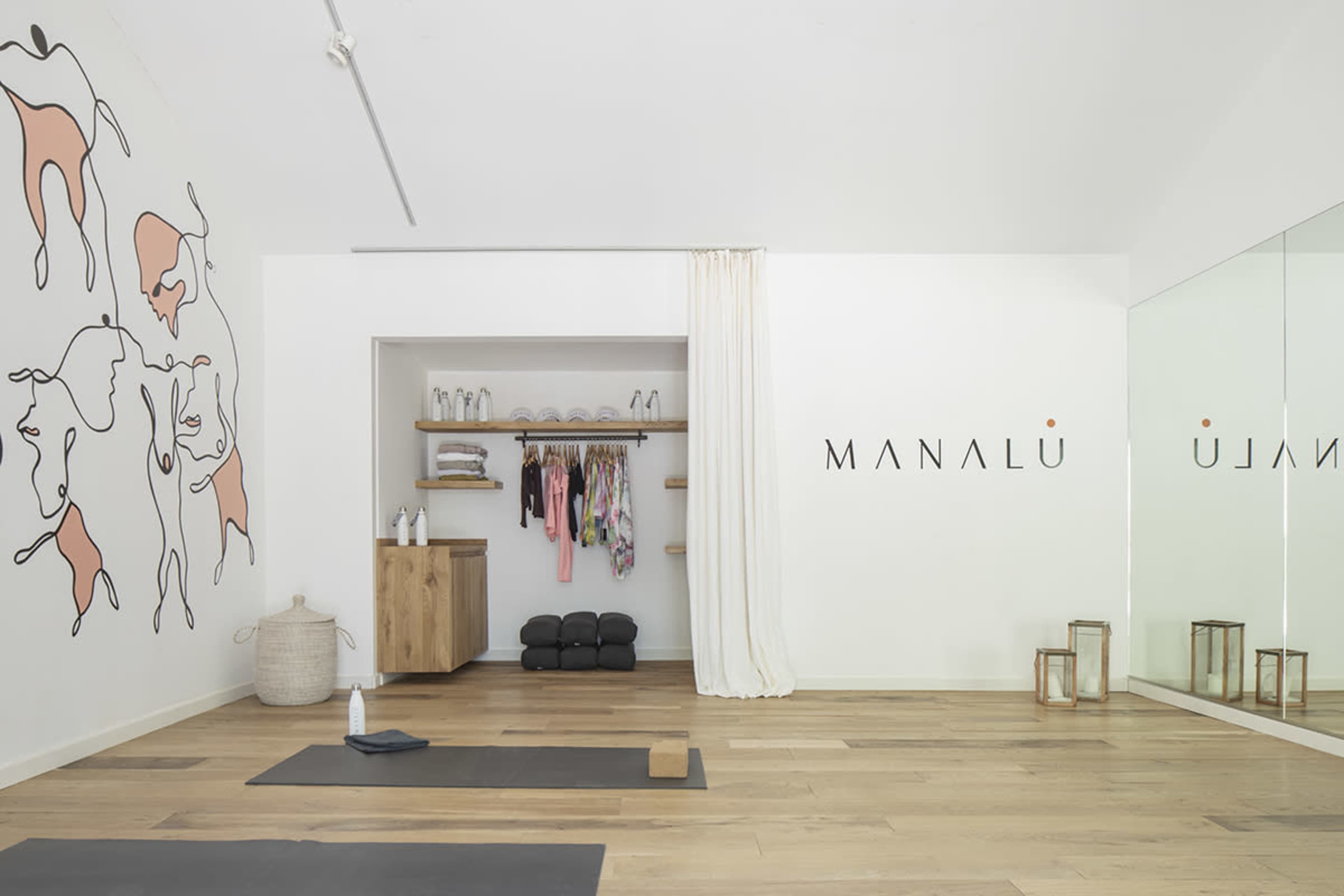 A minimalist yoga studio with a mirrored wall, wooden flooring, organized shelves of yoga attire, and two mats laid out on the ground.