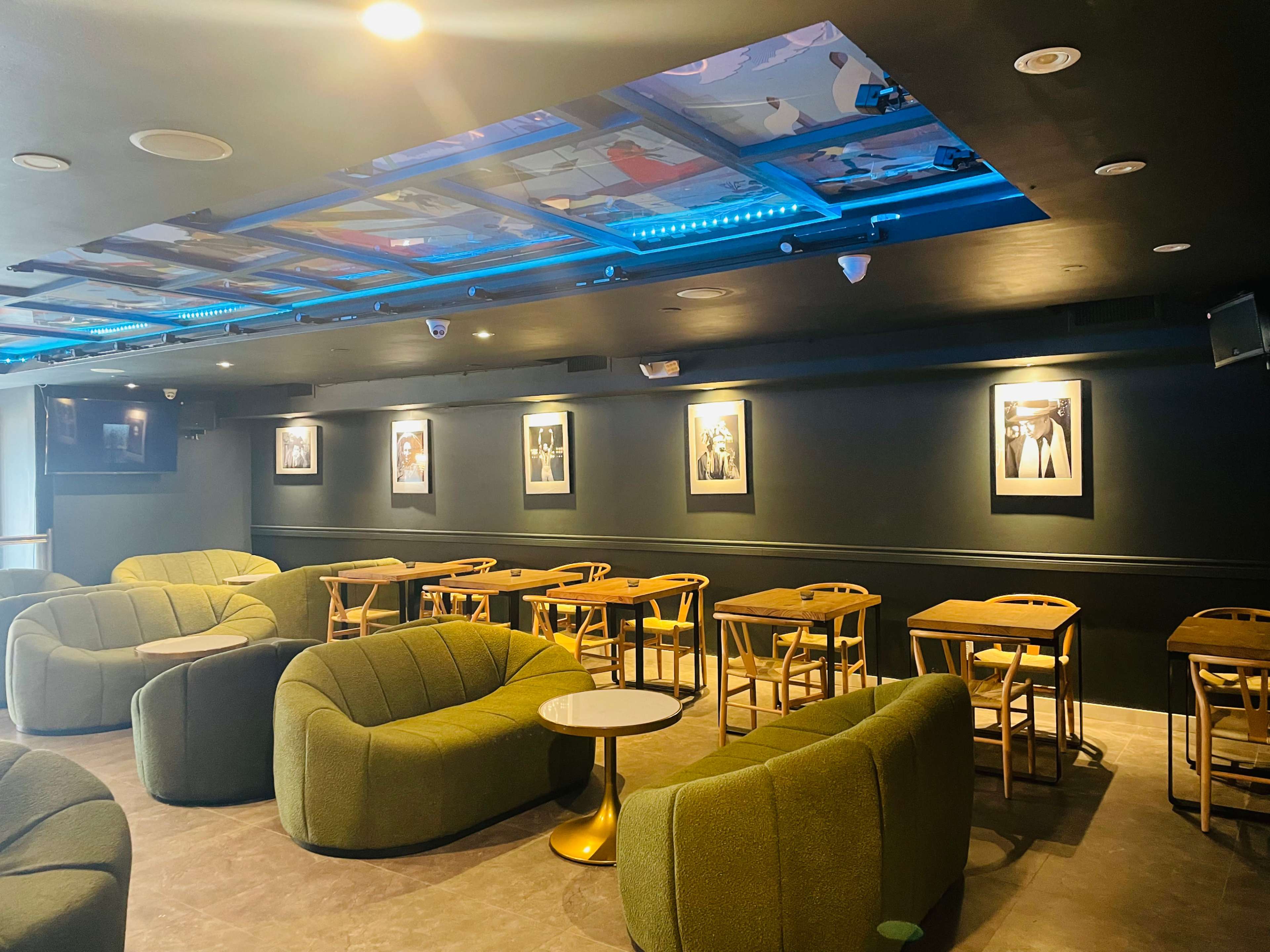 A modern lounge area with green upholstered seating, wooden tables, and framed artwork on the dark walls.
