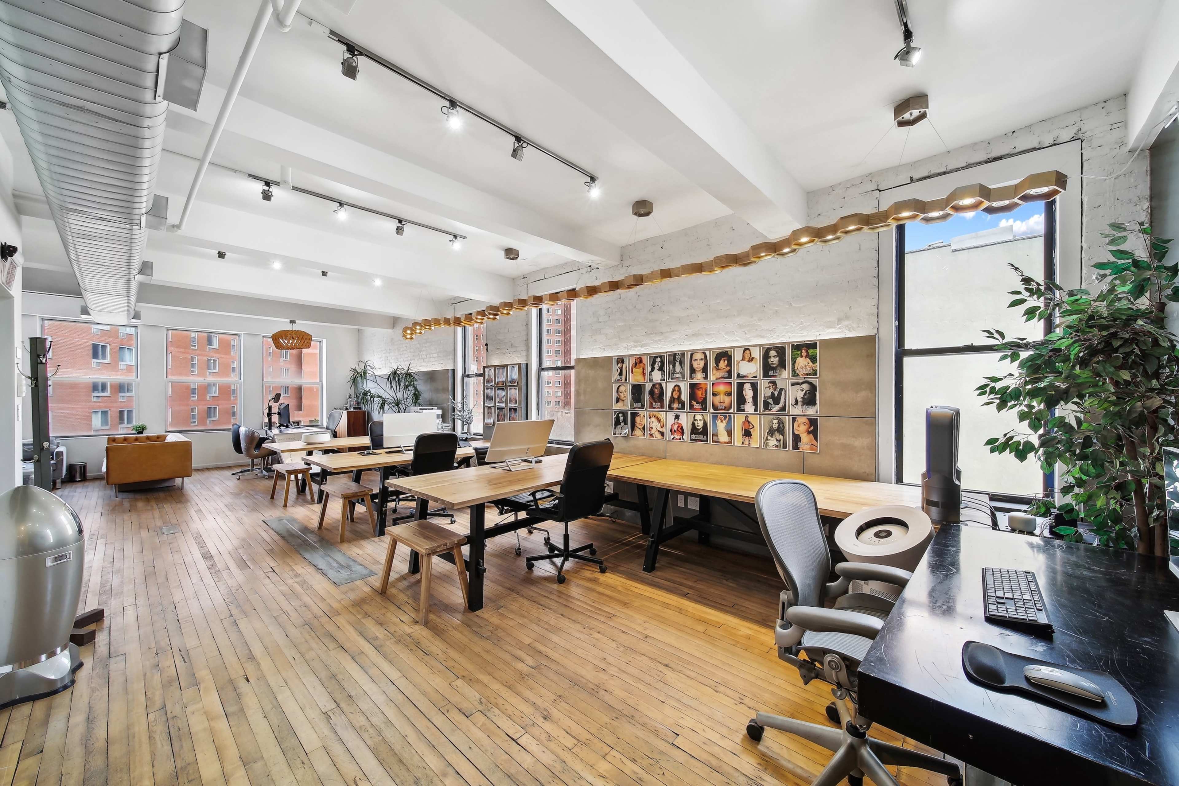 The image shows an open office space with wooden floors, several workstations, and a wall decorated with framed photos.