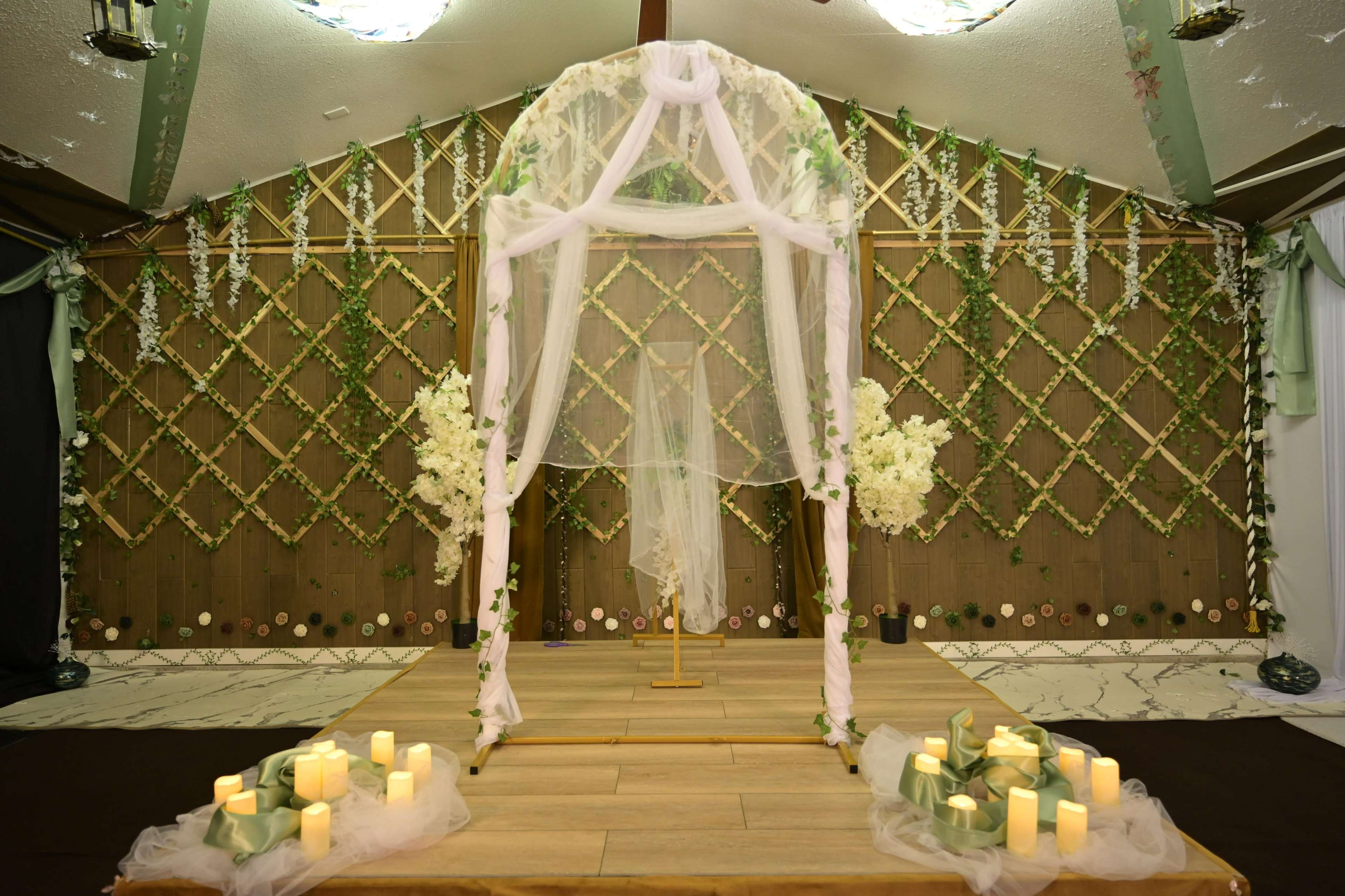 The image depicts a wedding altar adorned with white fabric, floral arrangements, and candles, set against a wooden backdrop with greenery accents.