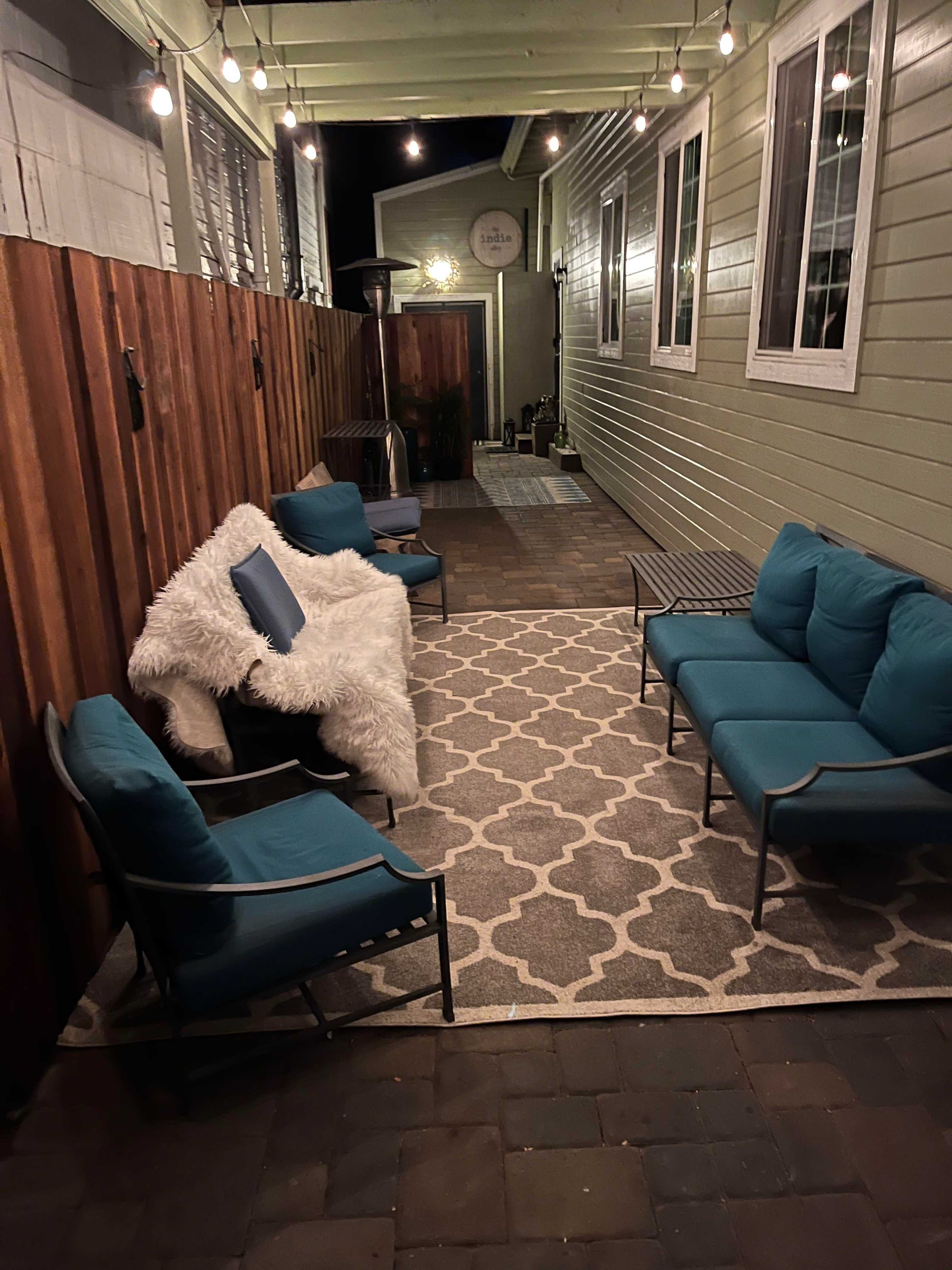 A well-lit outdoor patio area featuring turquoise seating and a textured rug, surrounded by wooden fencing and accented by string lights overhead.