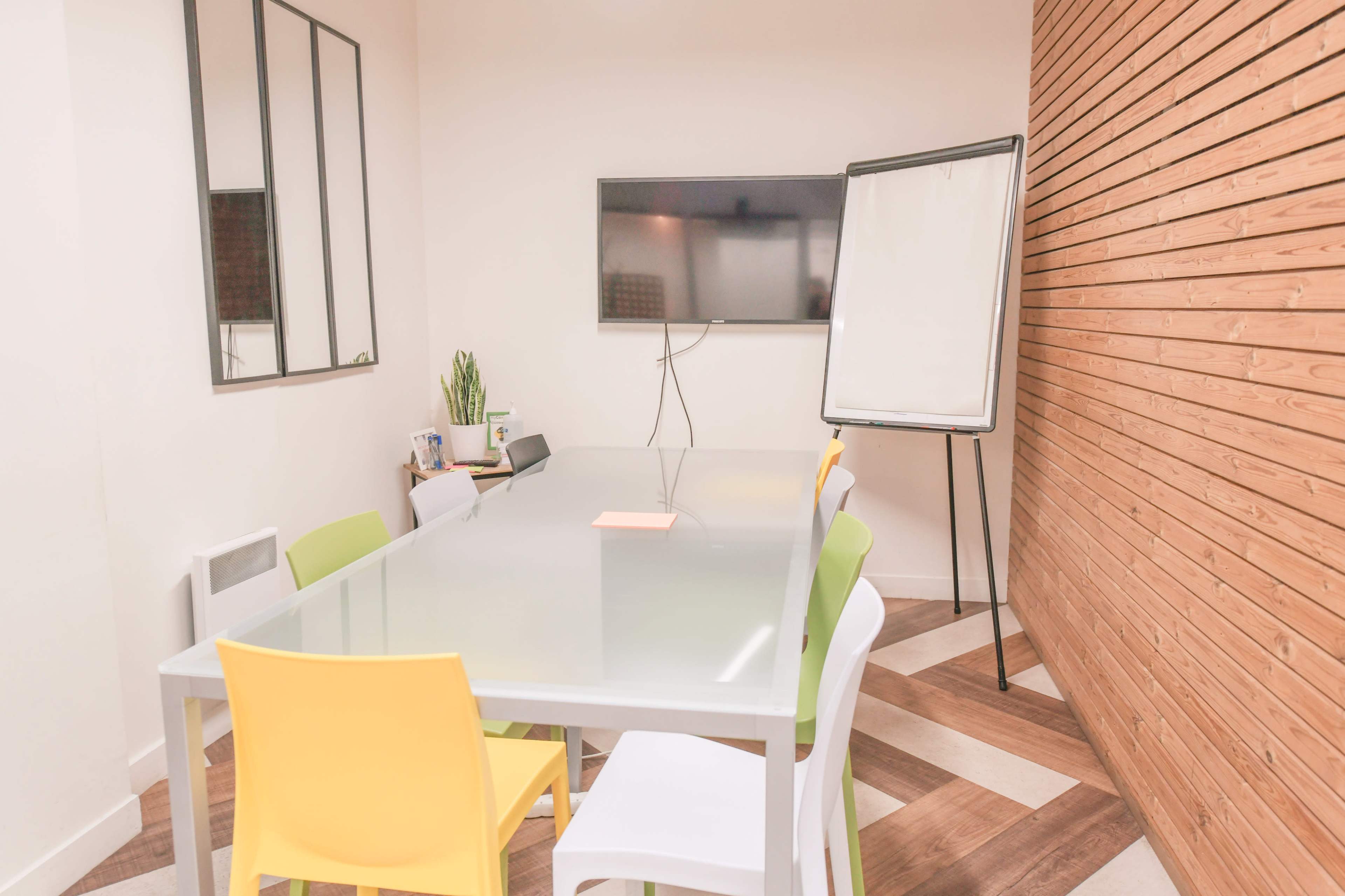 A conference room is set up with a glass table, colorful chairs, a wall-mounted television, and a flip chart on a stand.