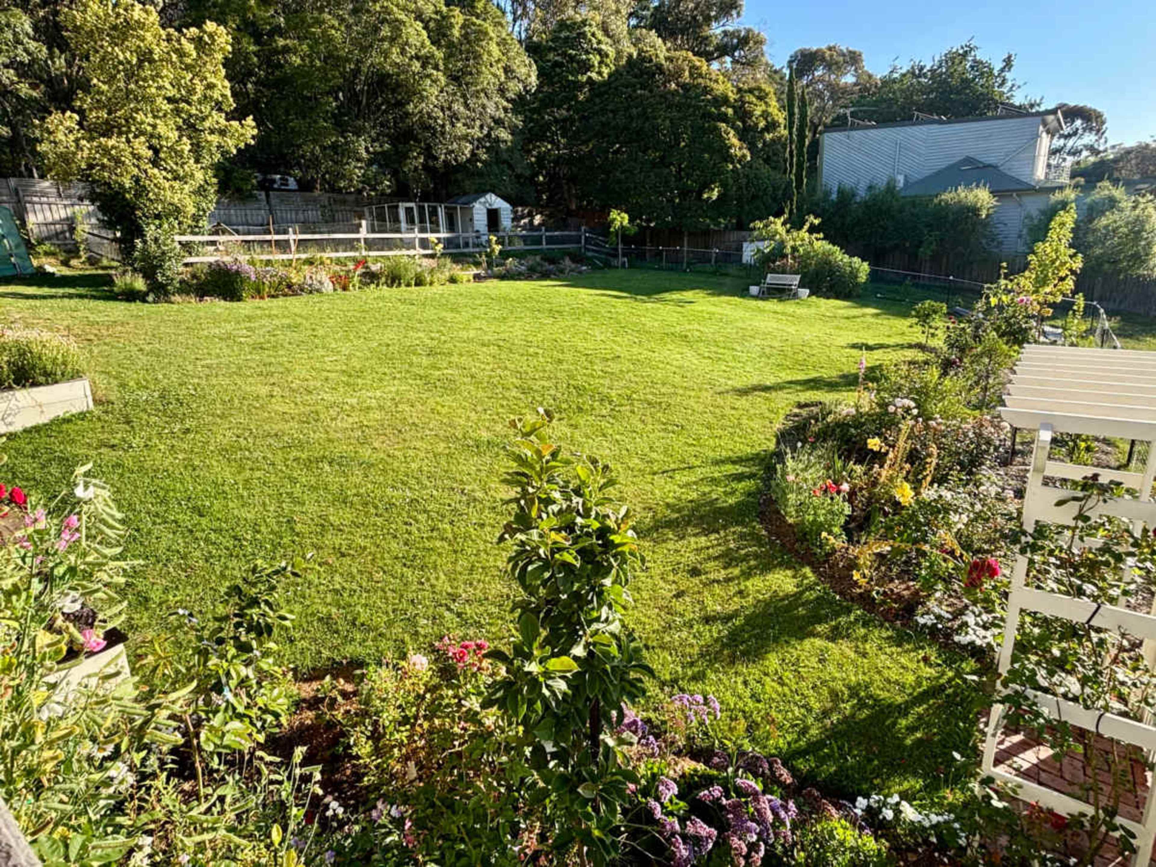 The image shows a well-maintained garden featuring a large grassy area surrounded by flower beds and a garden shed in the background.
