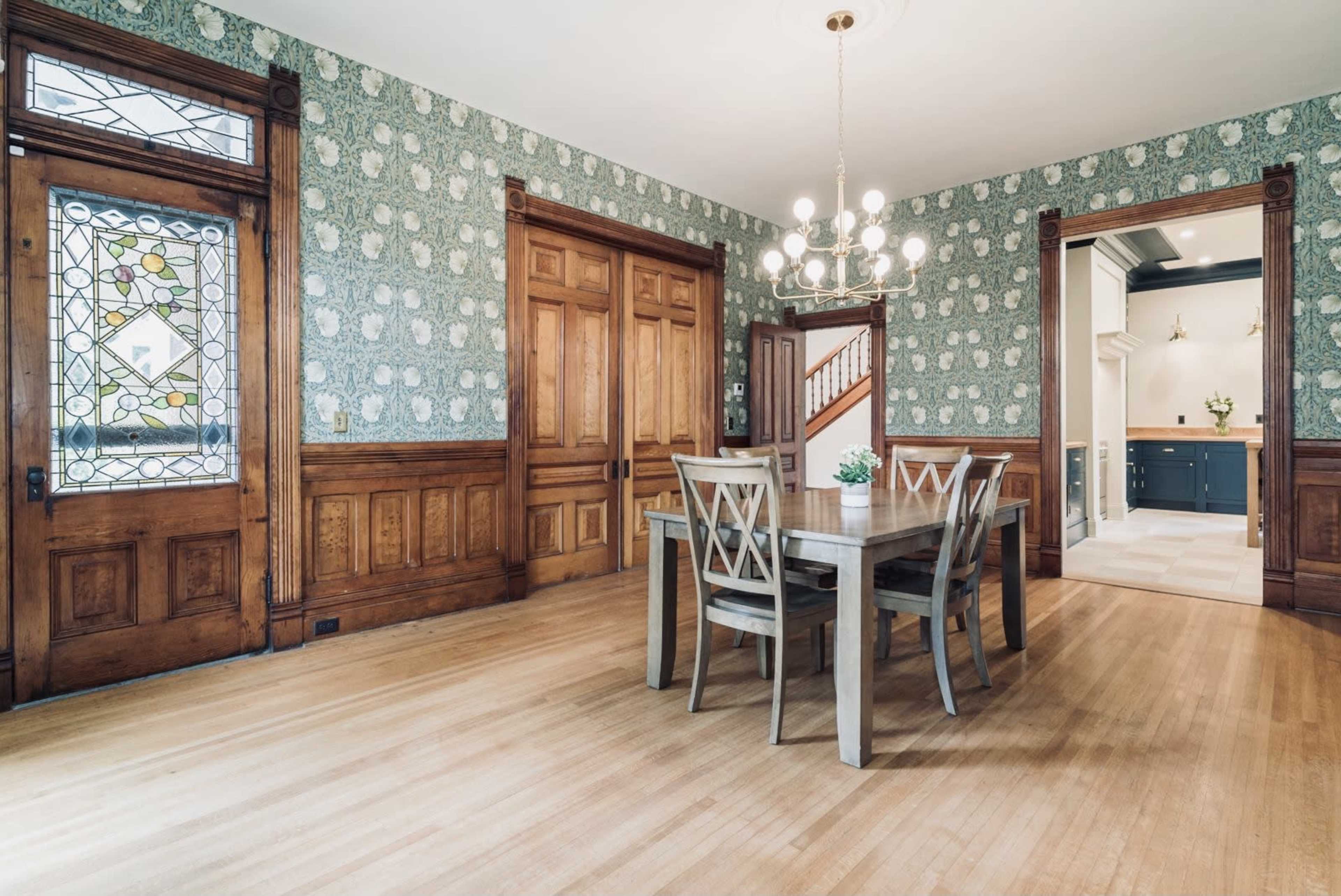 A dining room with a wooden table and chairs, surrounded by wallpaper featuring a floral design and framed by wooden doorways.