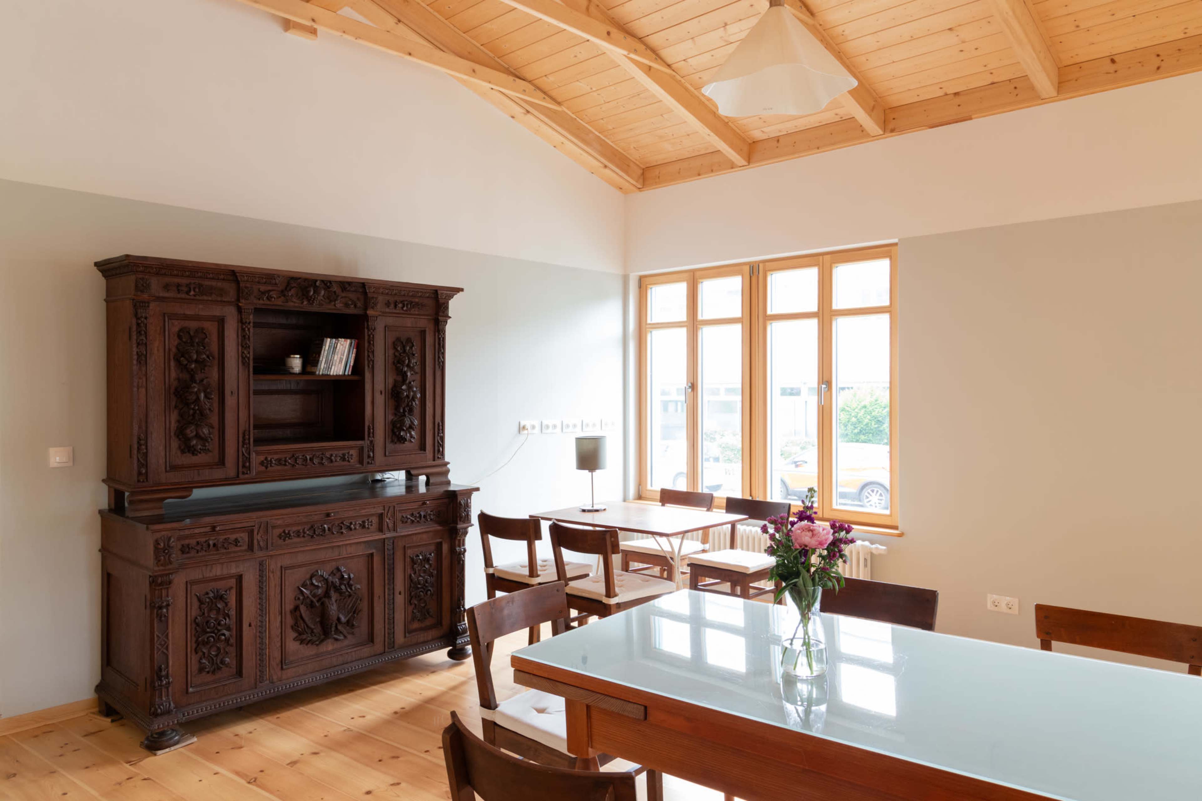 The image shows a spacious room with wooden beams, featuring a large antique wooden cabinet, a table set with chairs, and a vase of flowers on the table.