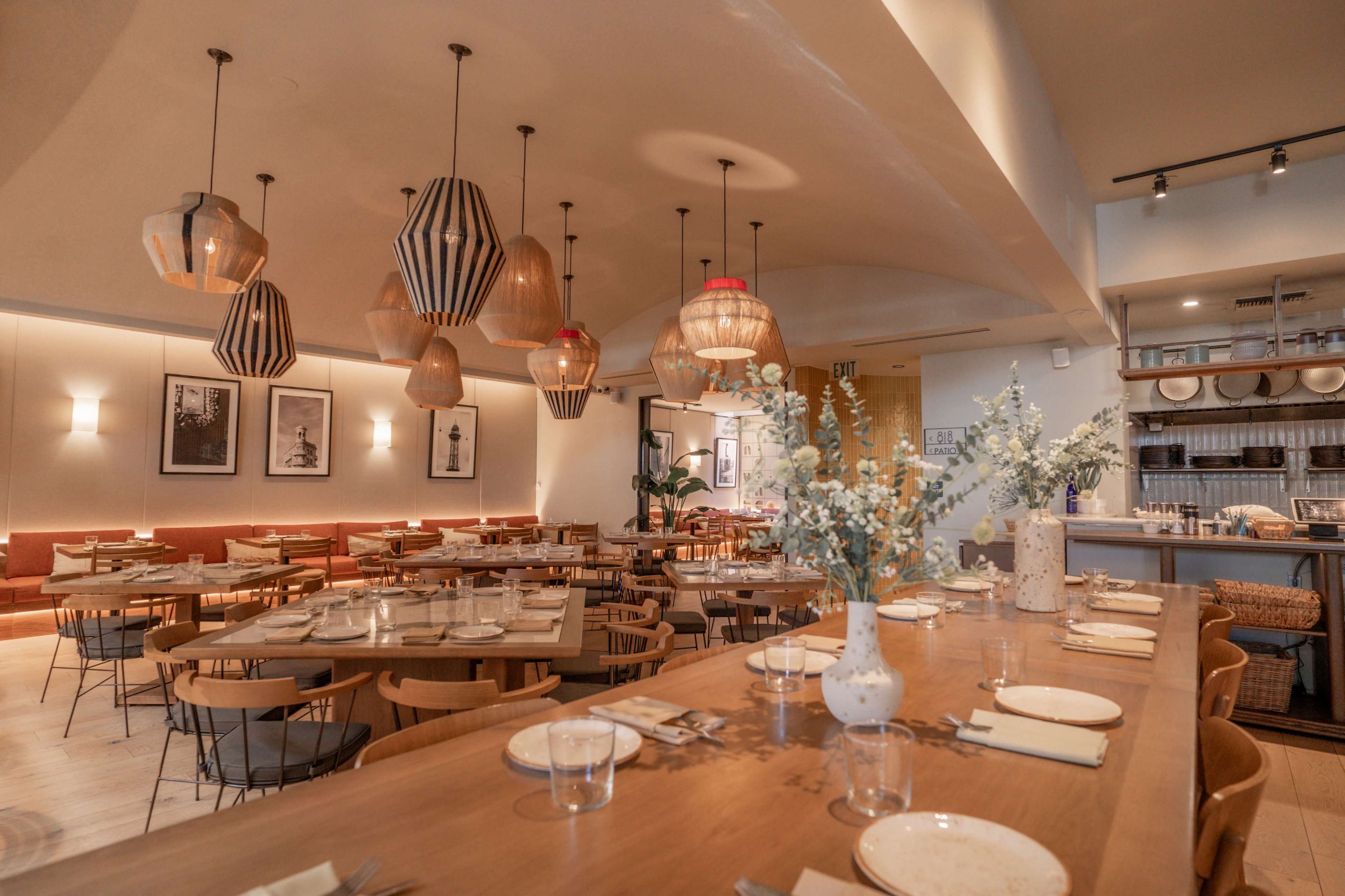The image shows a modern restaurant interior featuring wooden tables set for dining, contemporary light fixtures hanging from the ceiling, and decorative plants.