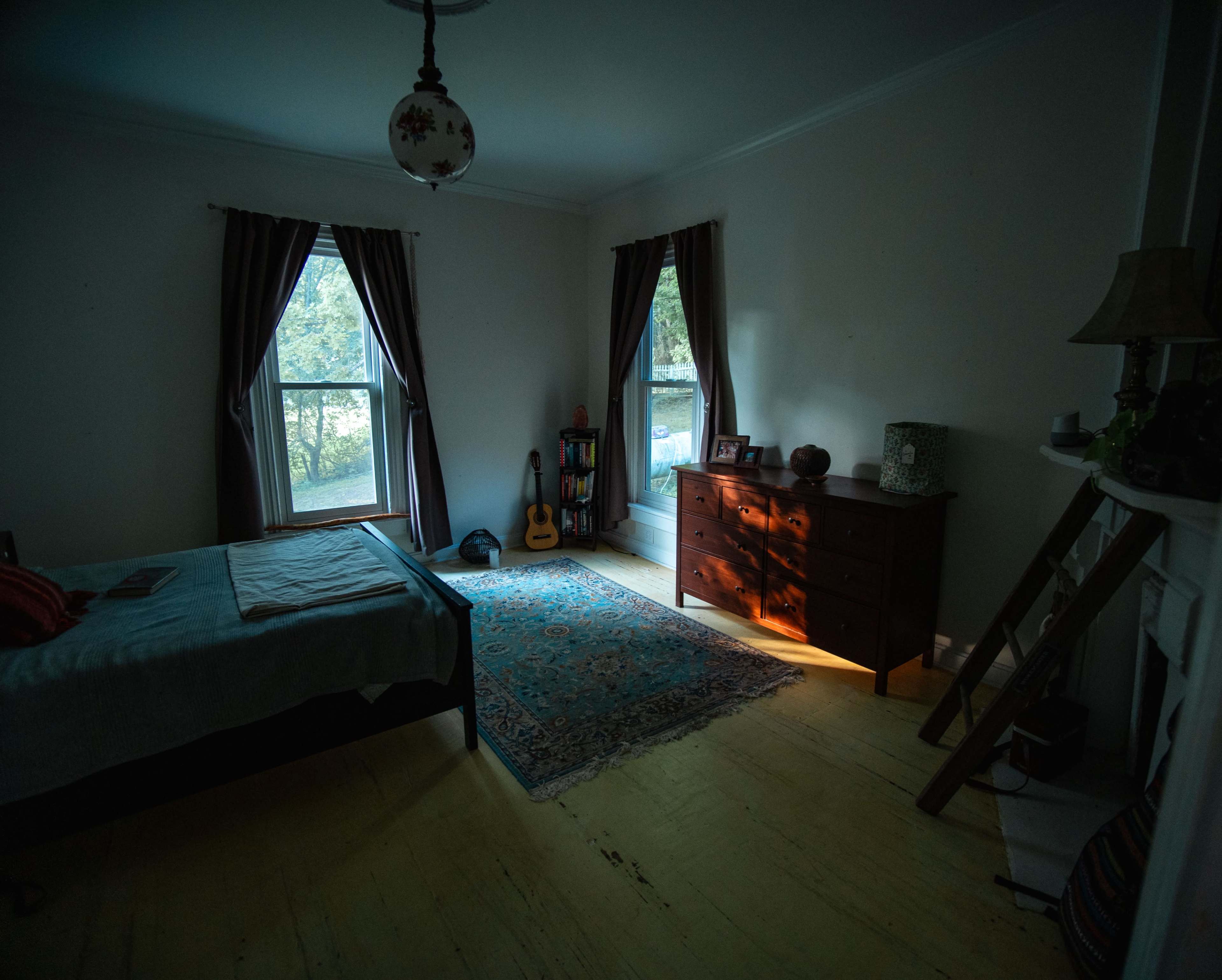A dimly lit bedroom features a bed, a dresser, and a guitar beside a window, with sunlight streaming through the drapes onto the floor.