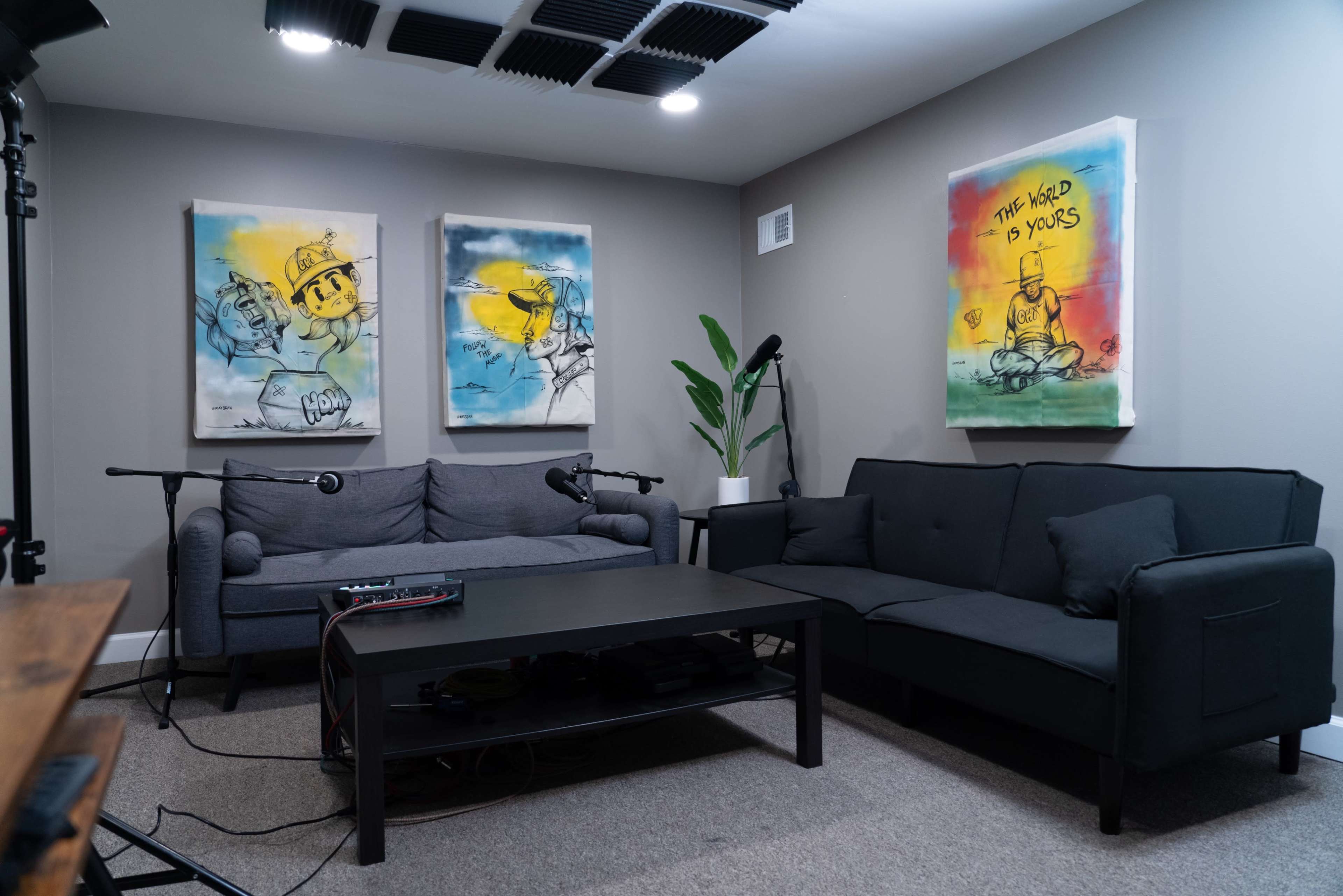 A cozy room features two sofas, a coffee table, and three vibrant abstract artworks on the walls, with microphones positioned for a recording setup.