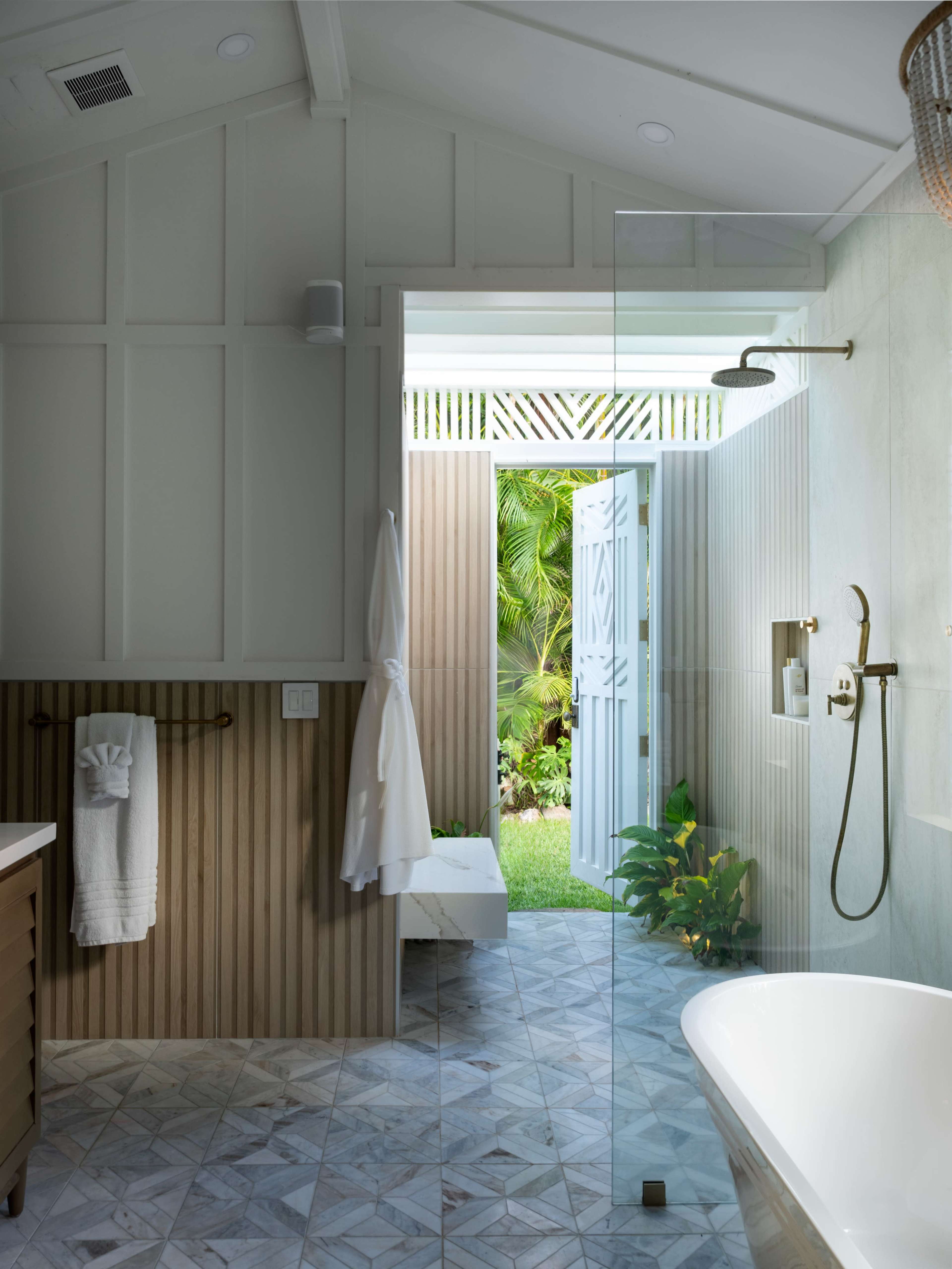 The image shows a modern bathroom featuring a glass shower enclosure, a freestanding bathtub, and a doorway leading to a lush garden.
