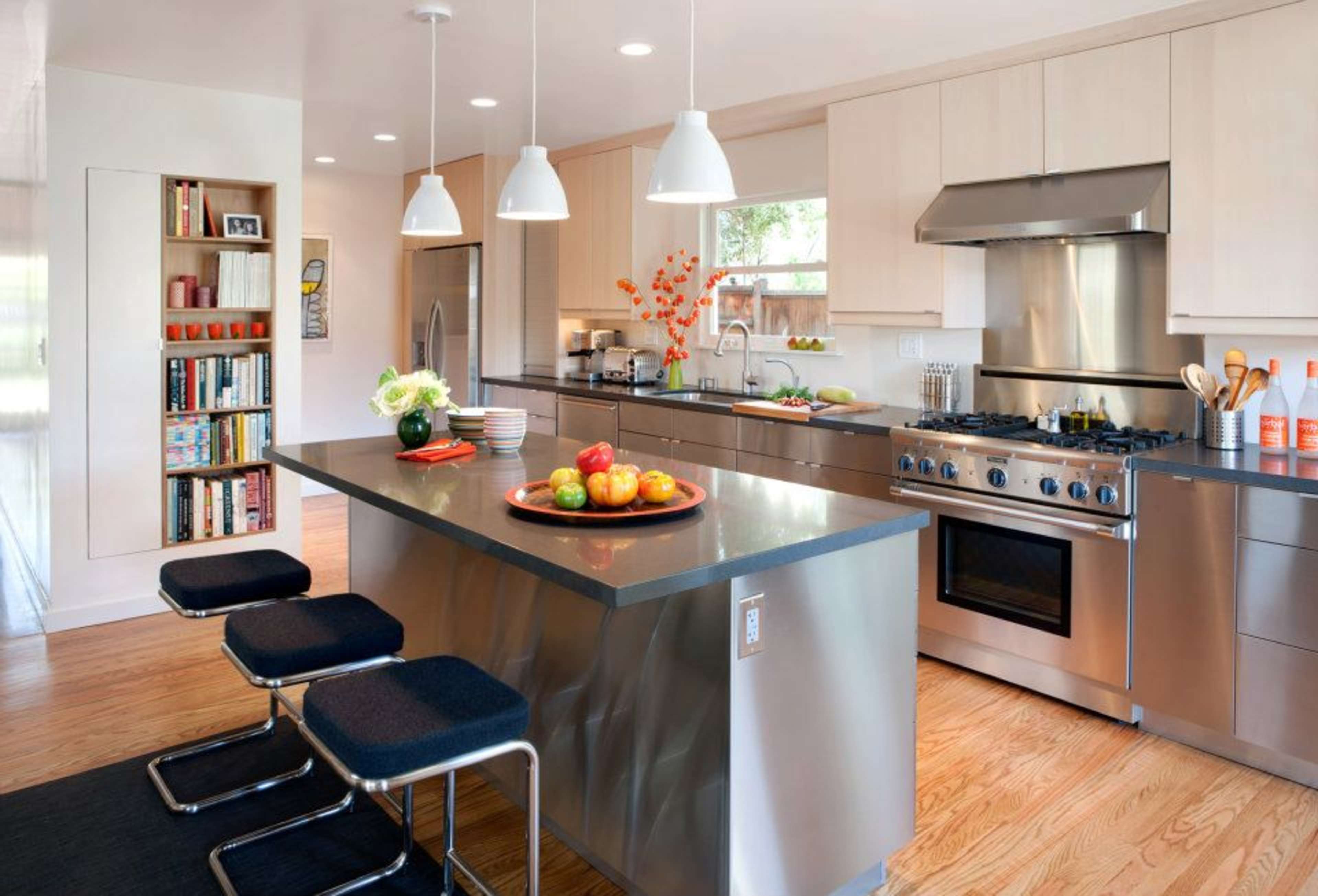 A modern kitchen features stainless steel appliances, a central island with a bowl of apples, and sleek cabinetry with a bookshelf along one wall.