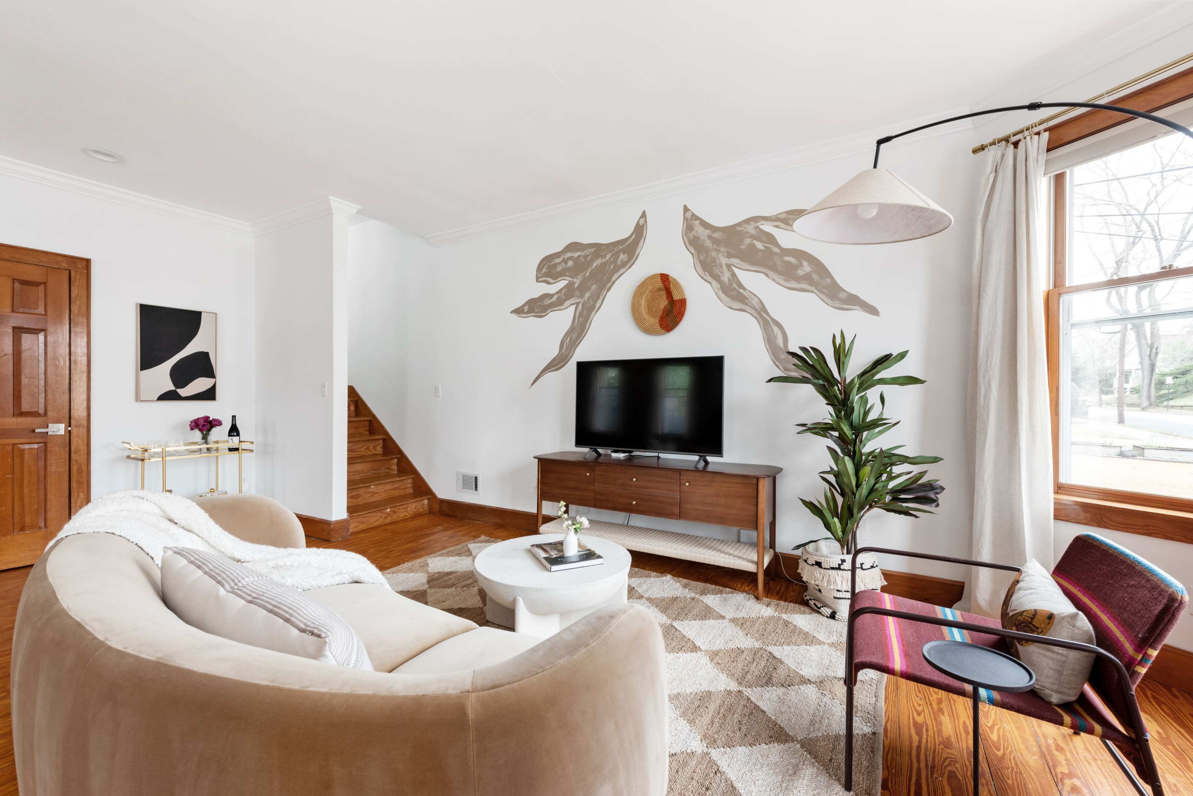 A cozy living room features a circular sofa, a flat-screen TV mounted on a wooden console, and a colorful accent chair beside a large window.