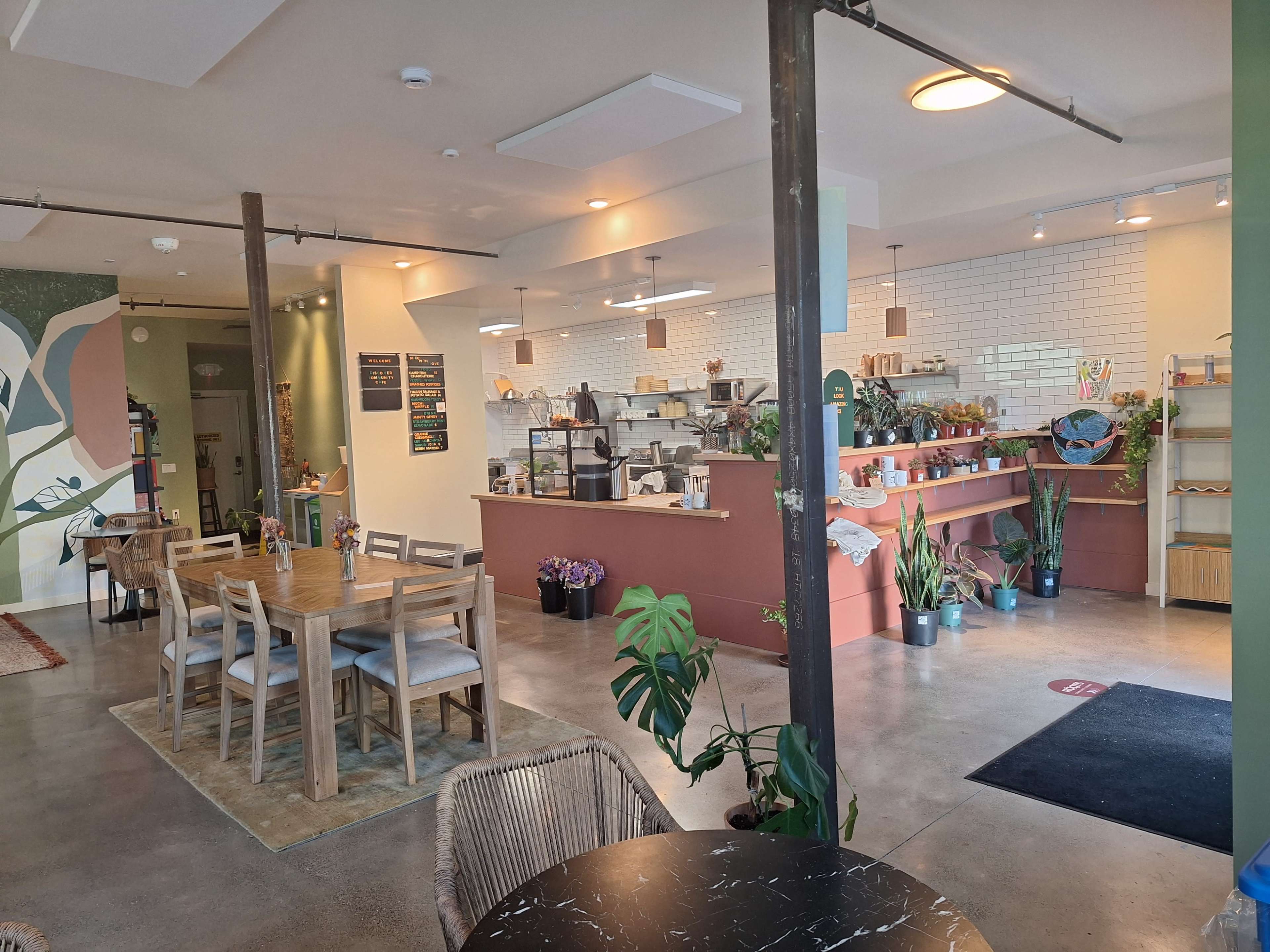 The image shows a modern café interior with a seating area, a service counter, and decorative plants.