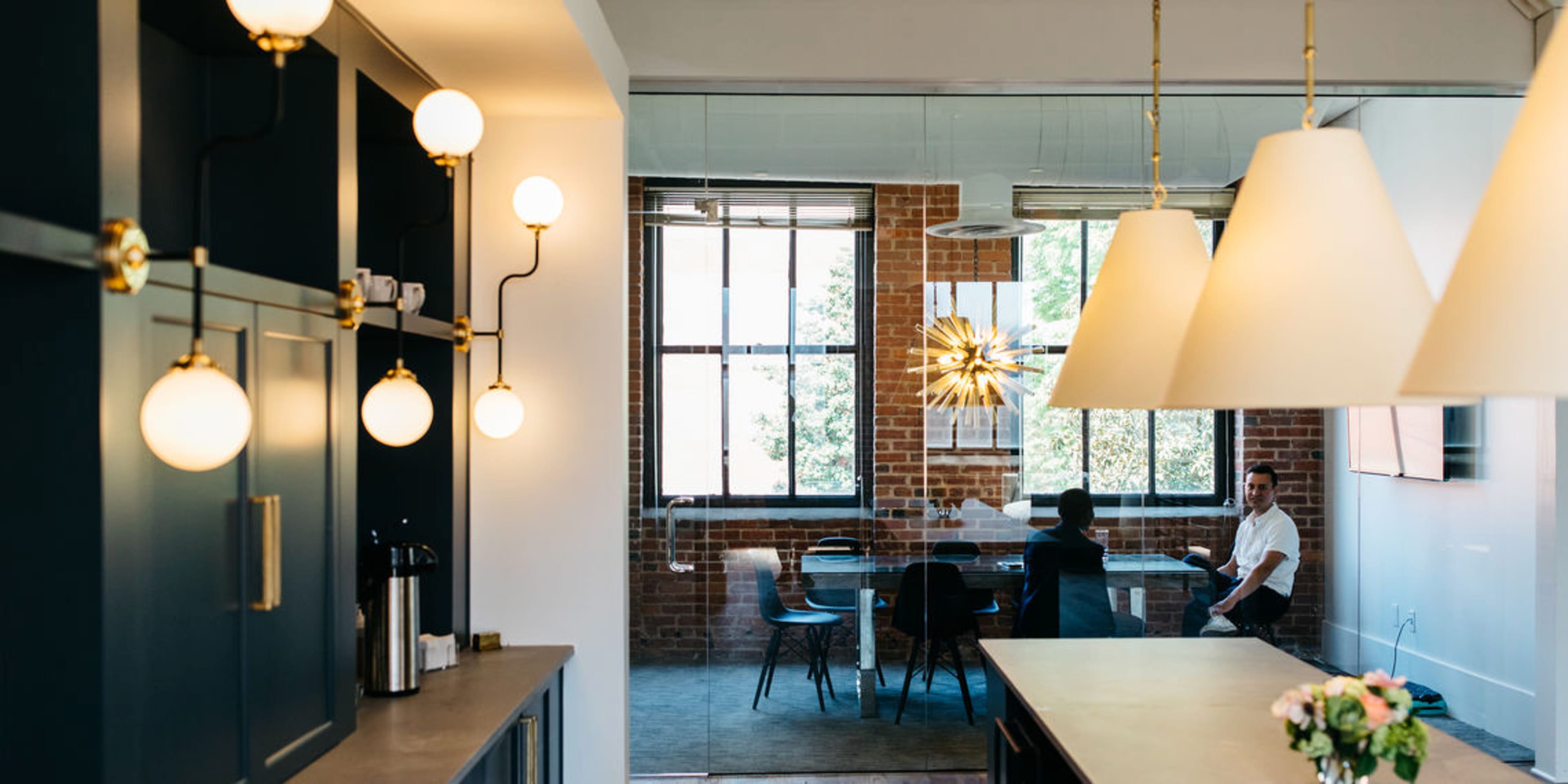 The image shows a modern workspace featuring a kitchenette area with pendant lights, and a glass-walled meeting room where two people are engaged in a discussion.