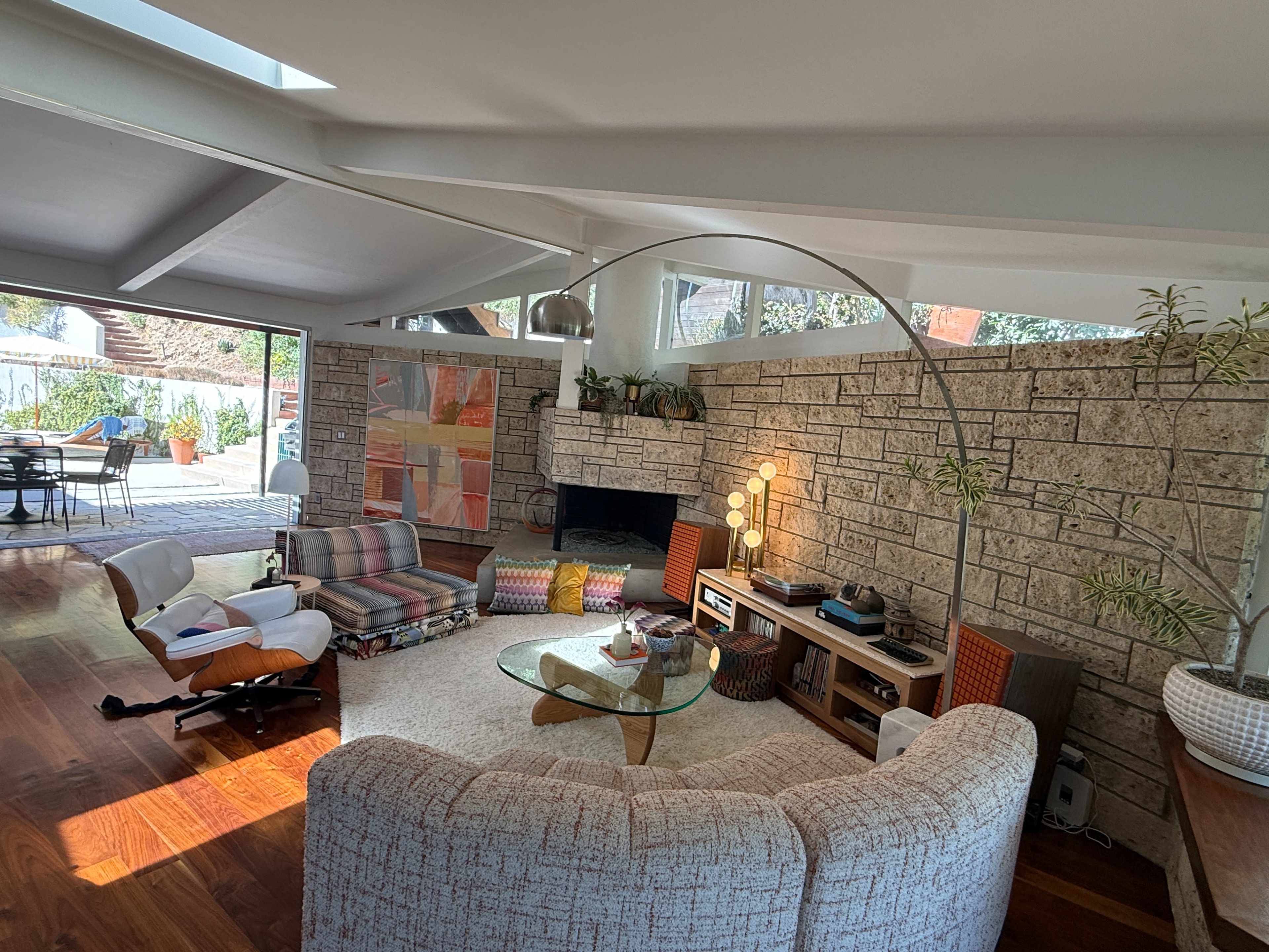 The image shows a modern living room with a curved seating area, a large window providing natural light, and a stone wall featuring decorative elements.