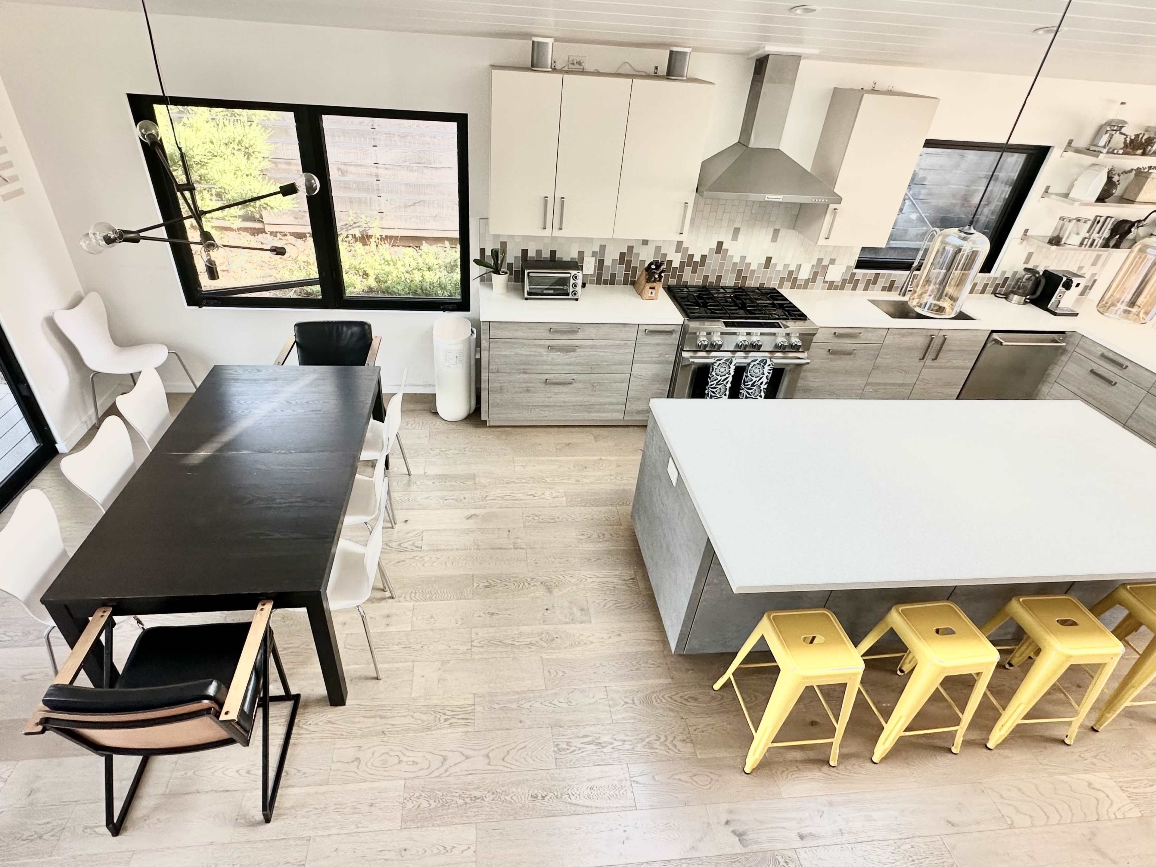 A modern kitchen features a central island with yellow stools, a dining table with black chairs, and large windows allowing natural light to fill the space.