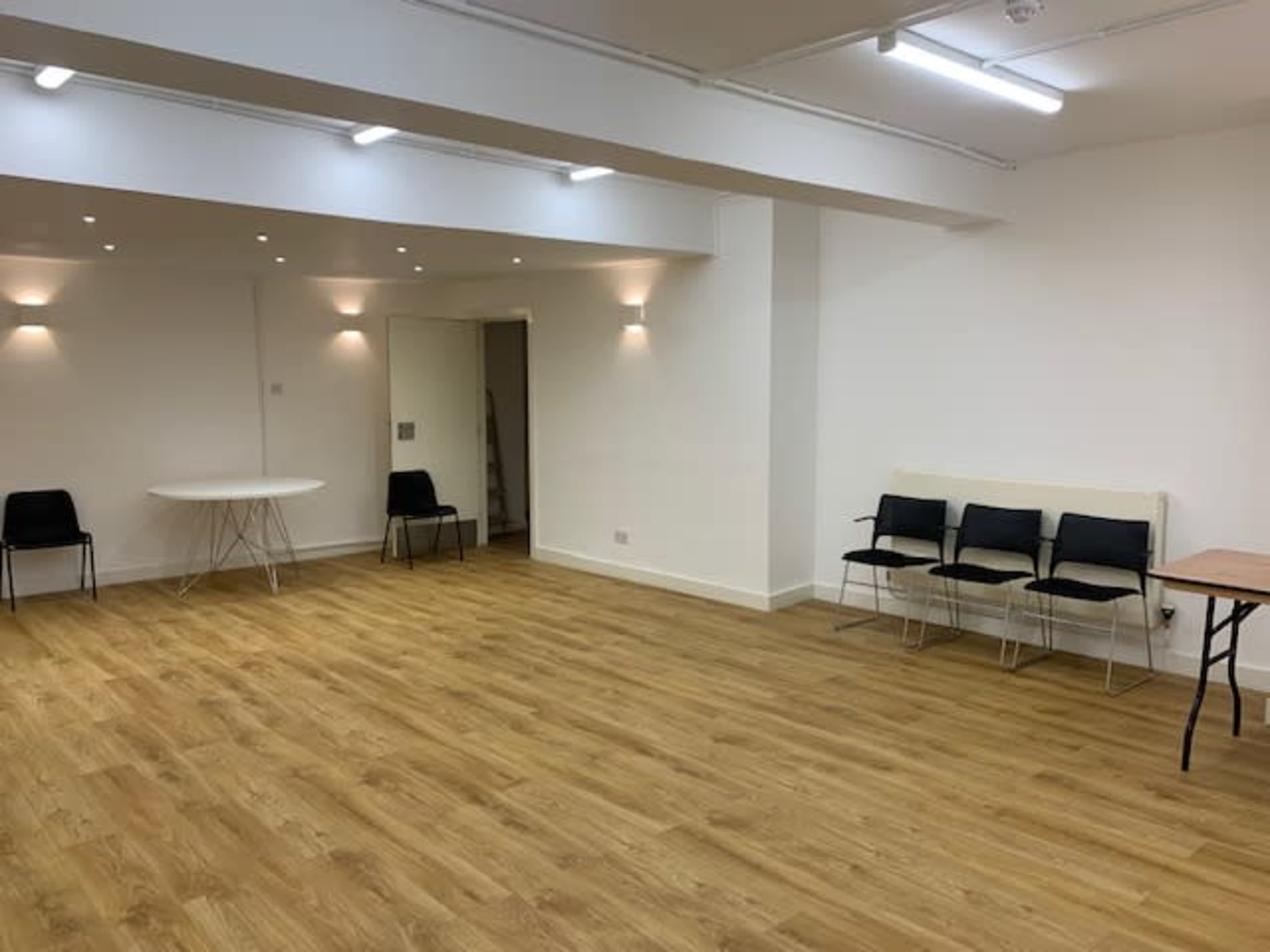 A spacious room with wooden flooring, featuring a round white table, several black chairs, and soft wall lighting.