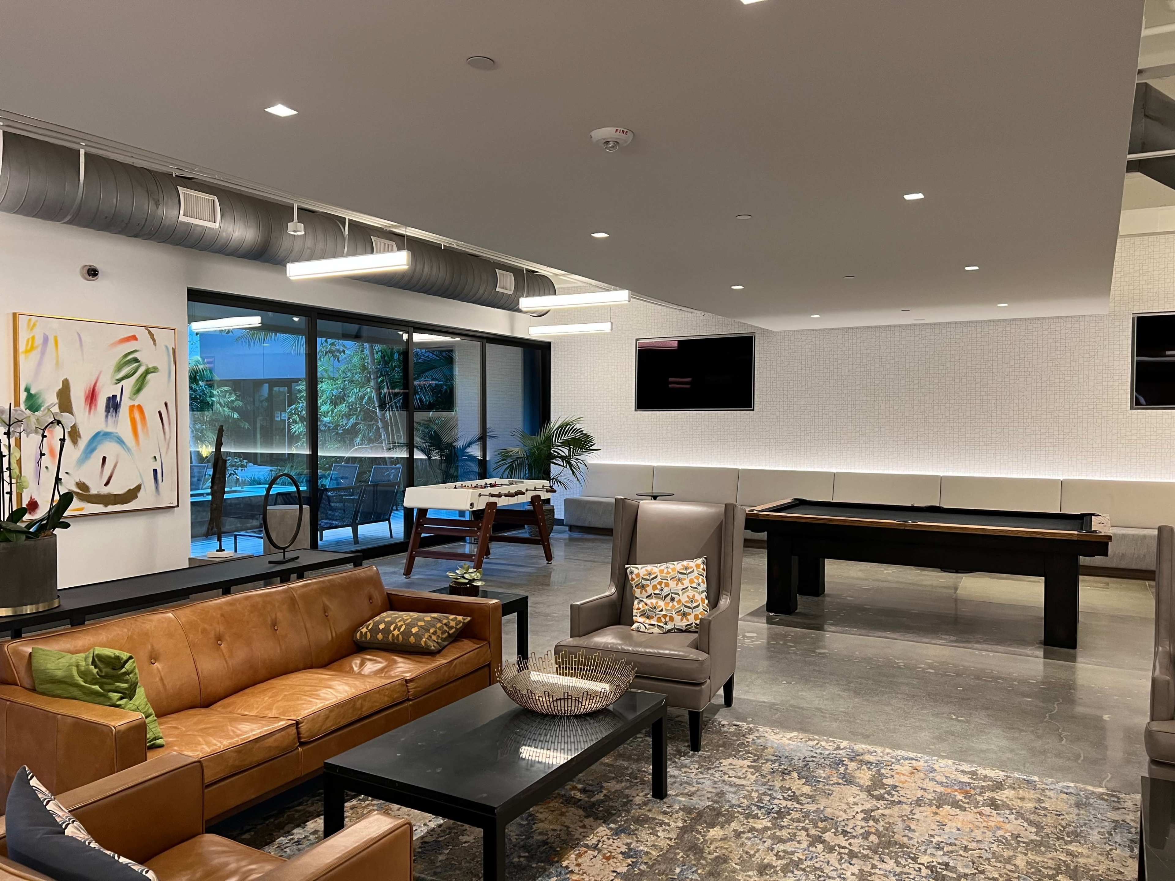 The image shows a modern lounge area with leather seating, a pool table, and large windows overlooking an outdoor space.