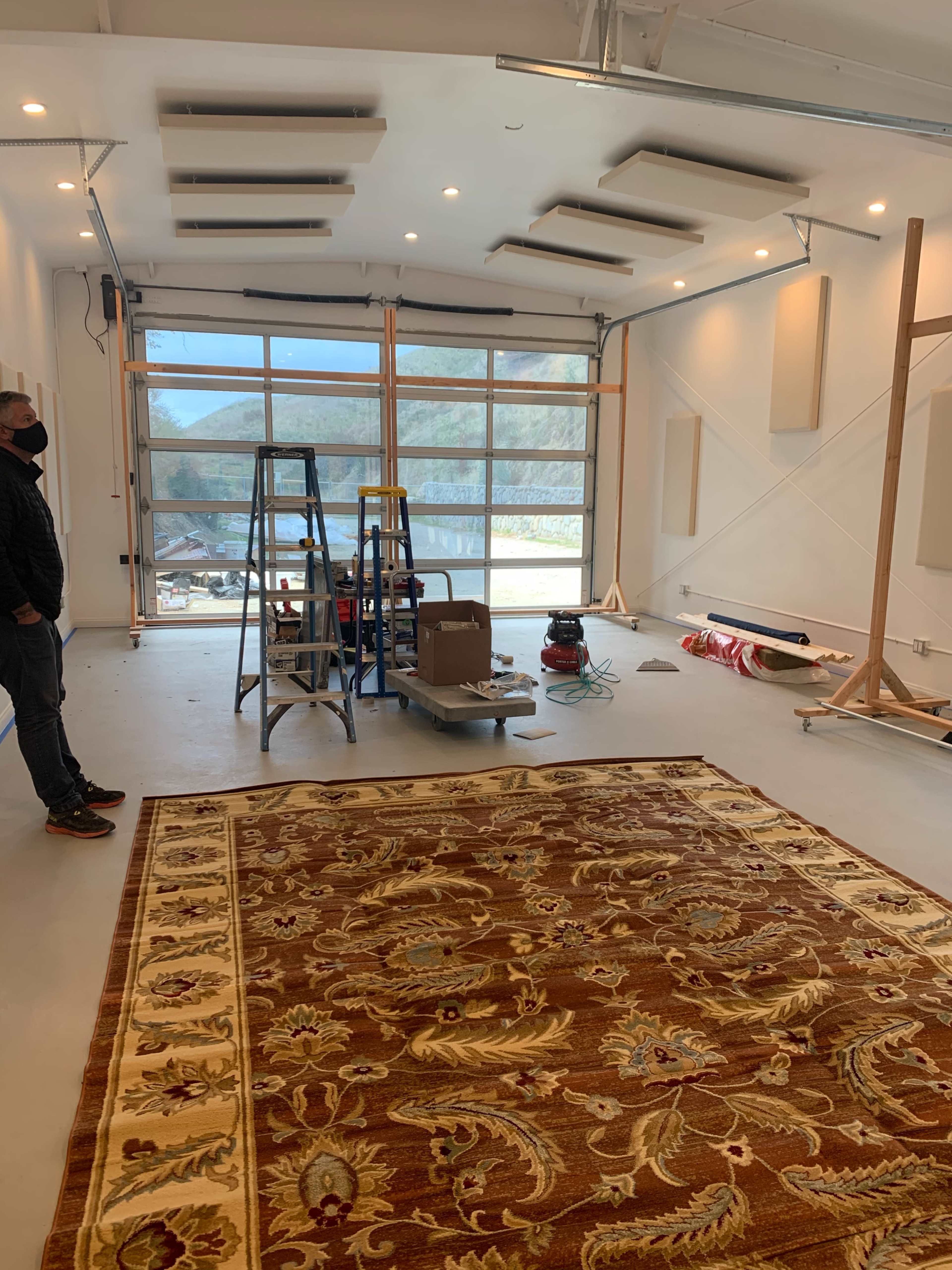 A spacious room is under construction, featuring a large decorative rug on the floor and various tools and equipment, including ladders and a vacuum, set against a backdrop of large windows.