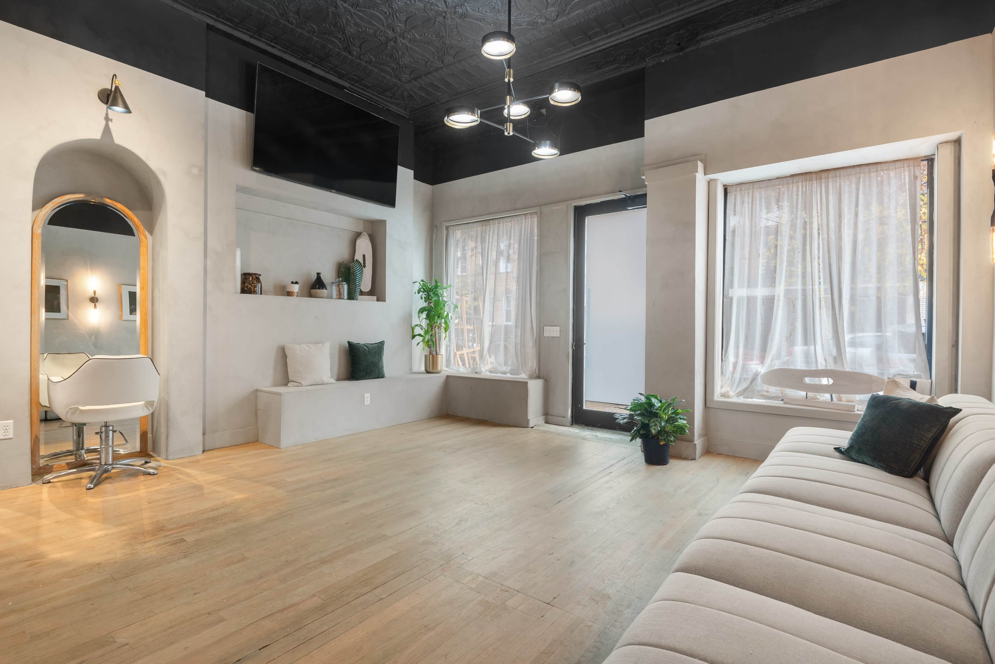 The image shows a minimalist interior space featuring light-colored walls, wooden flooring, a large window with sheer curtains, a sofa, and a chair in a corner.
