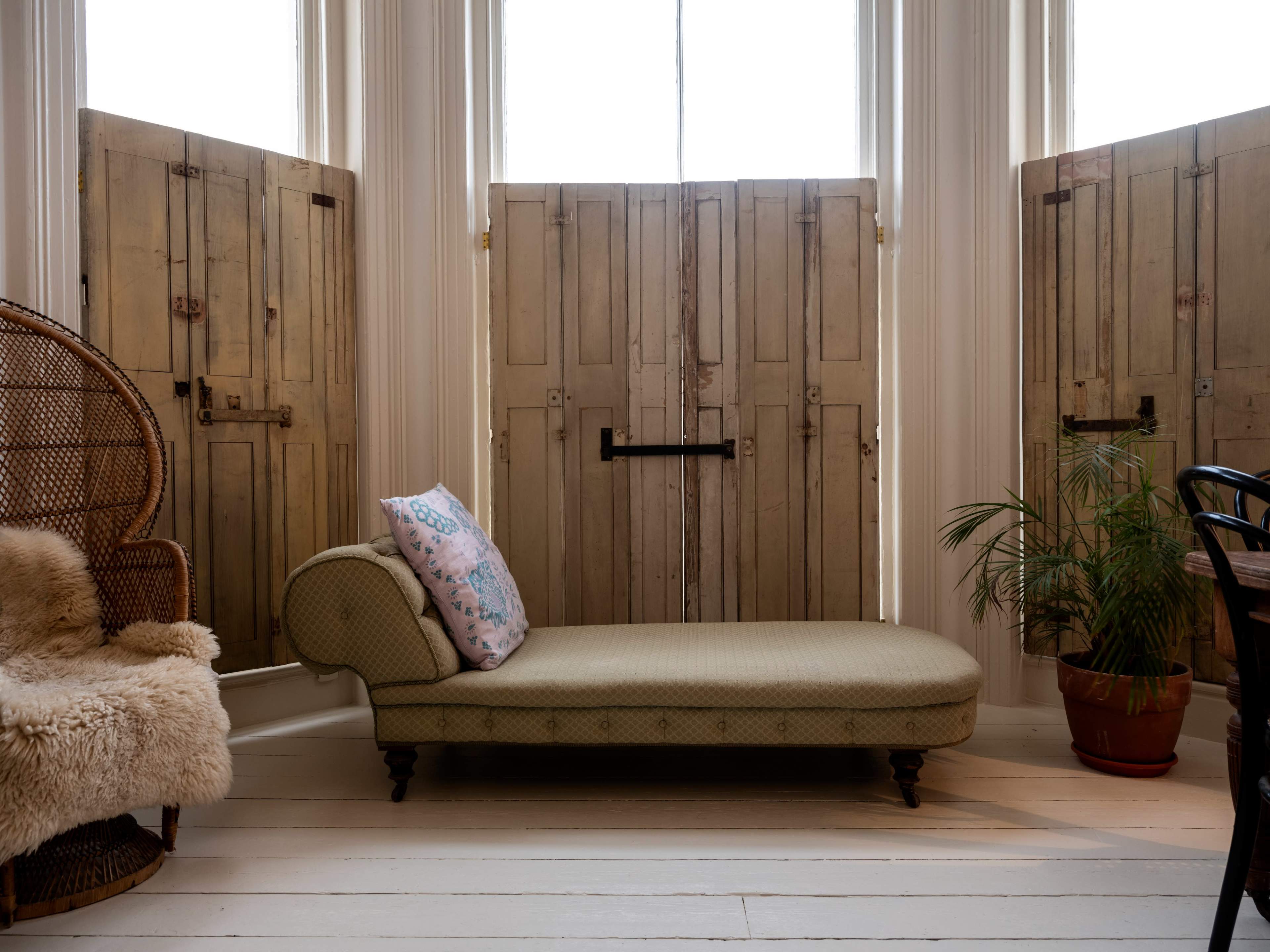A vintage green chaise lounge placed between two wooden folding doors in a well-lit room with a potted plant.
