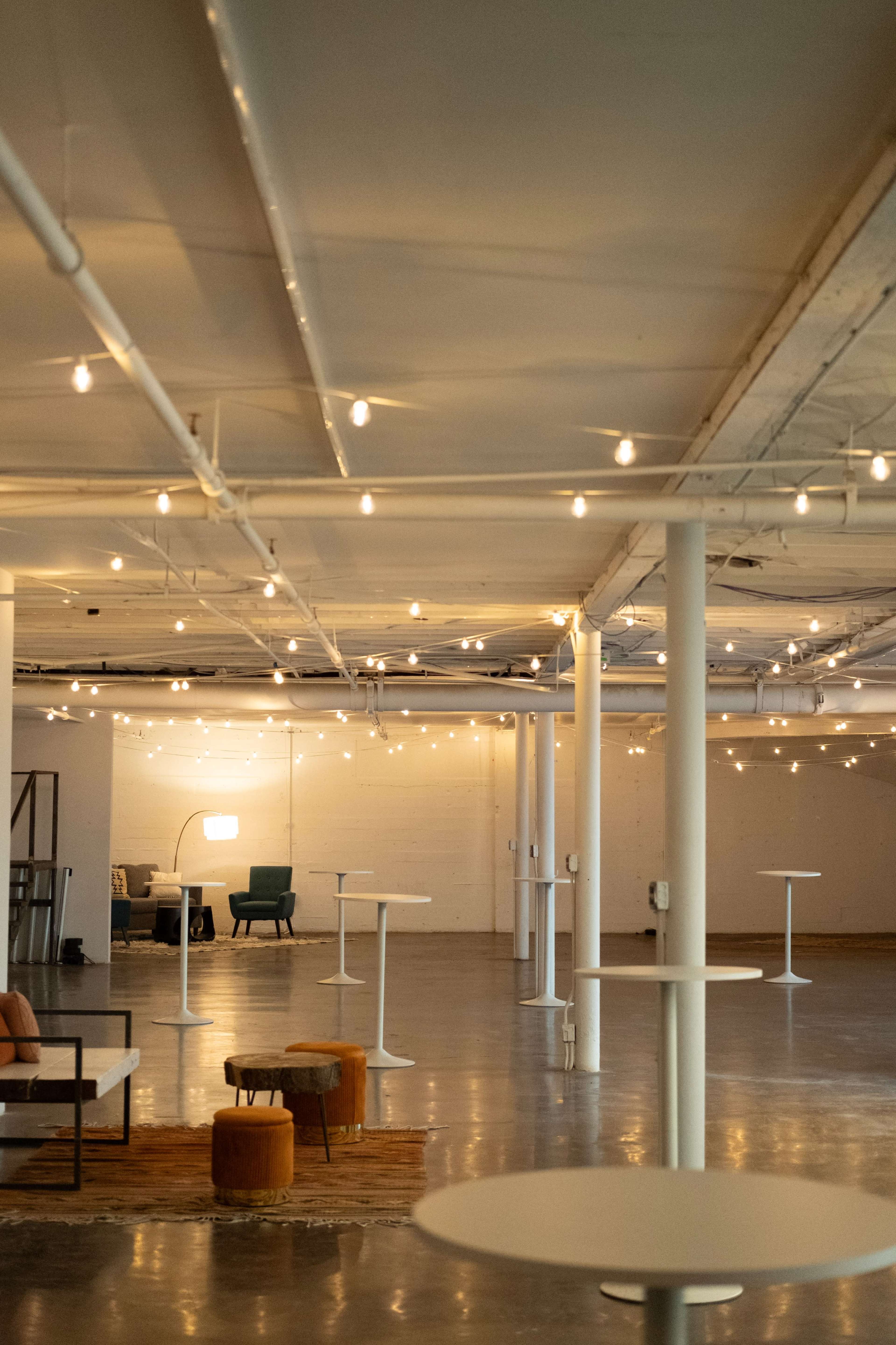 The image shows a spacious interior room with a polished concrete floor, several white tables, and soft lighting from string lights above.