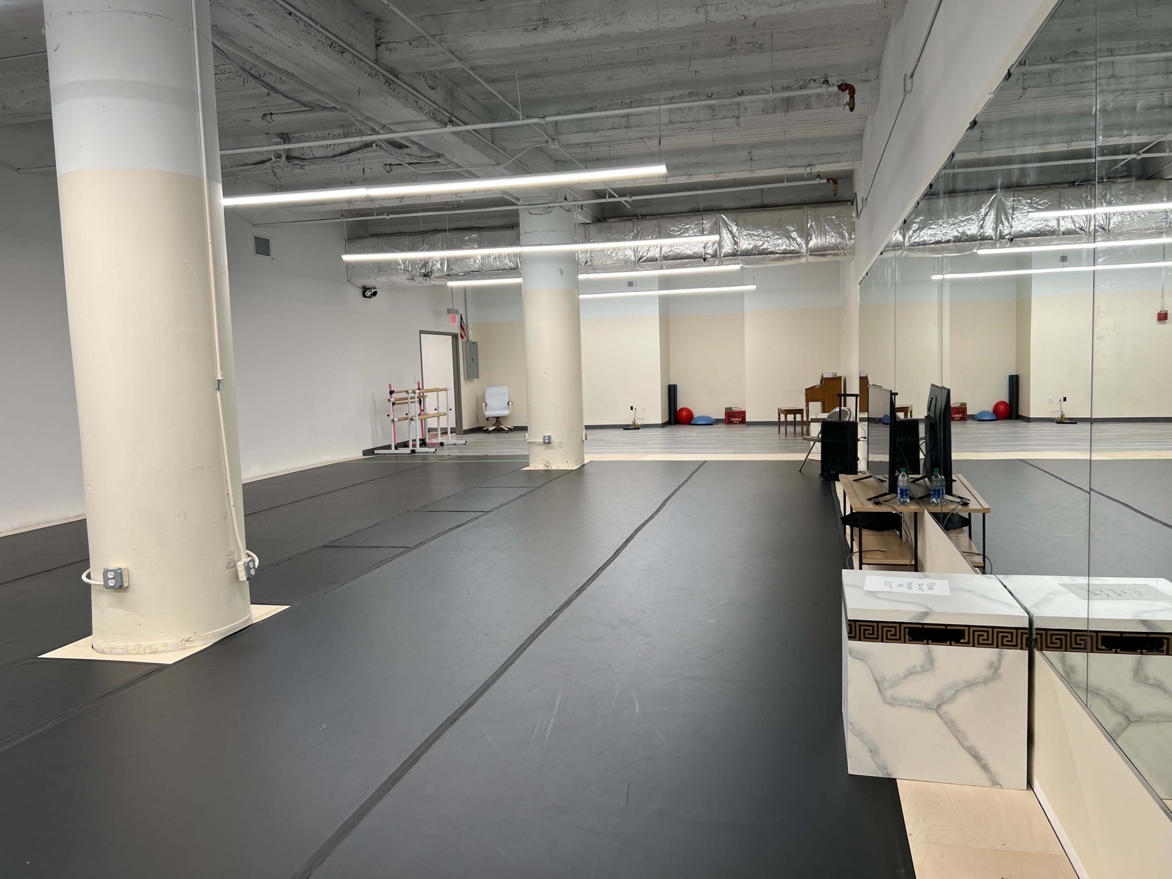 An empty gymnasium or studio with black mats, large columns, and mirrors along one wall.