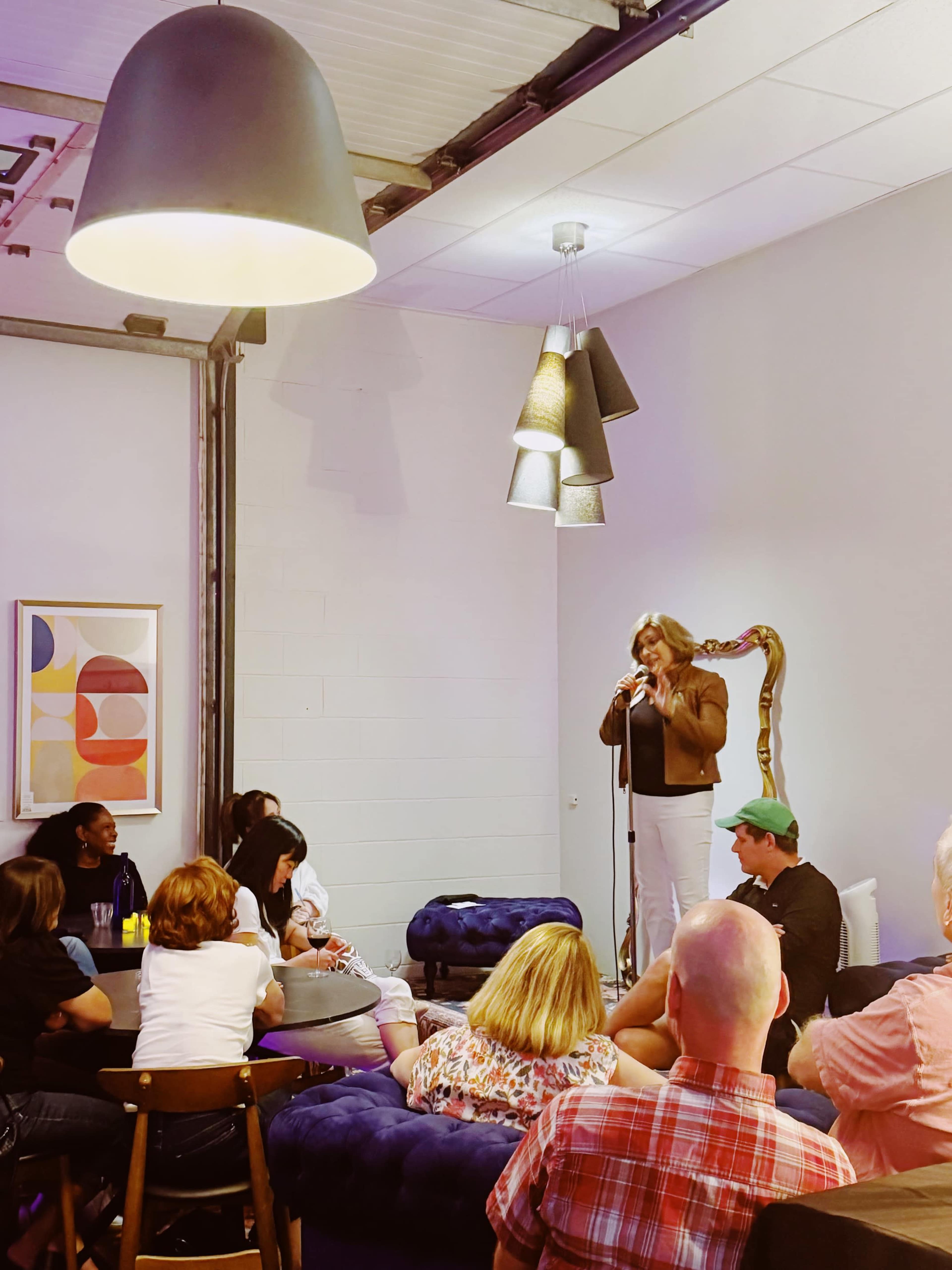 A woman stands on a small stage with a microphone, addressing an audience seated on couches and chairs in a well-lit room decorated with contemporary art.