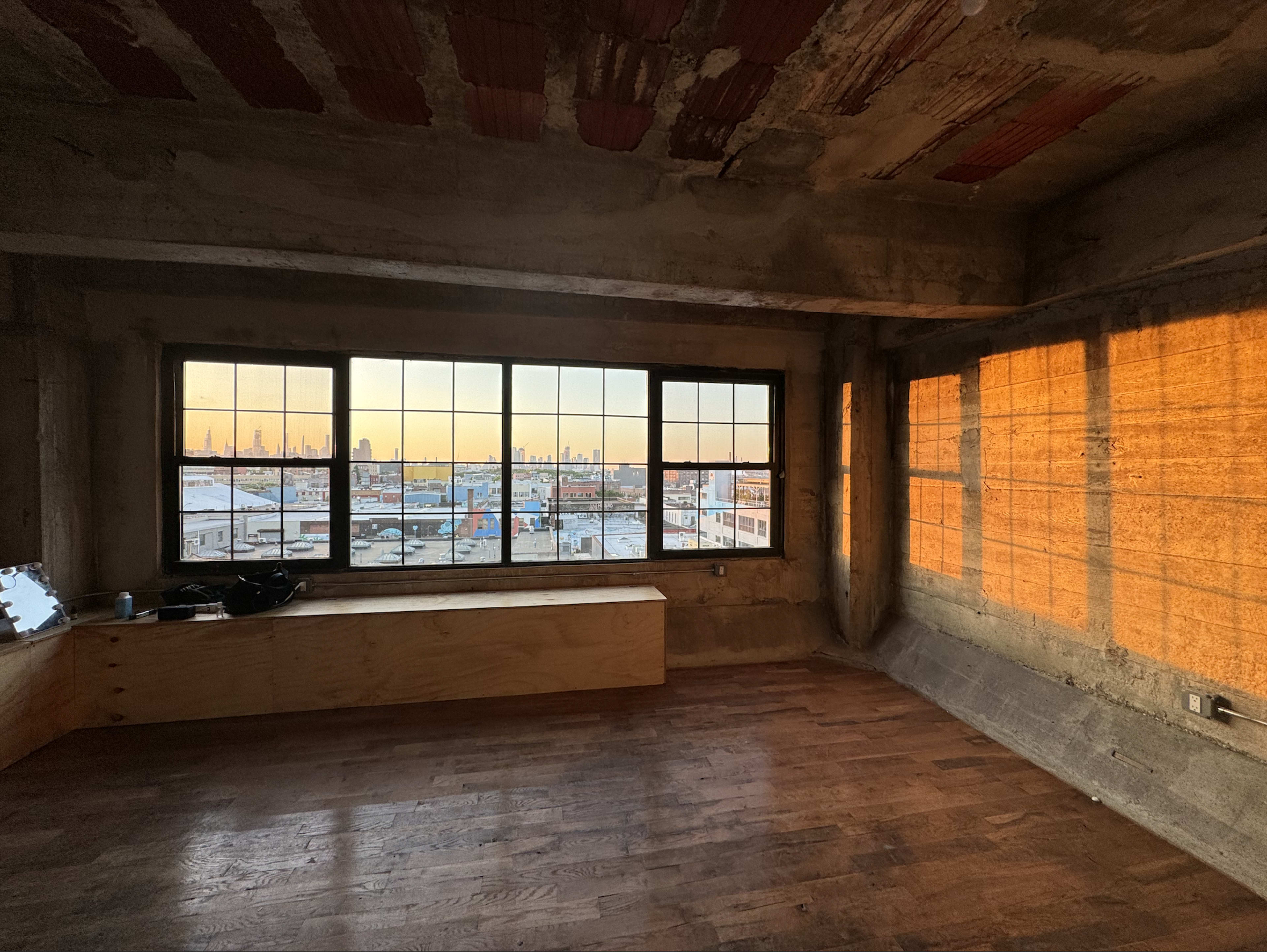 An unfinished interior room with large windows, allowing a view of a city skyline at sunset.