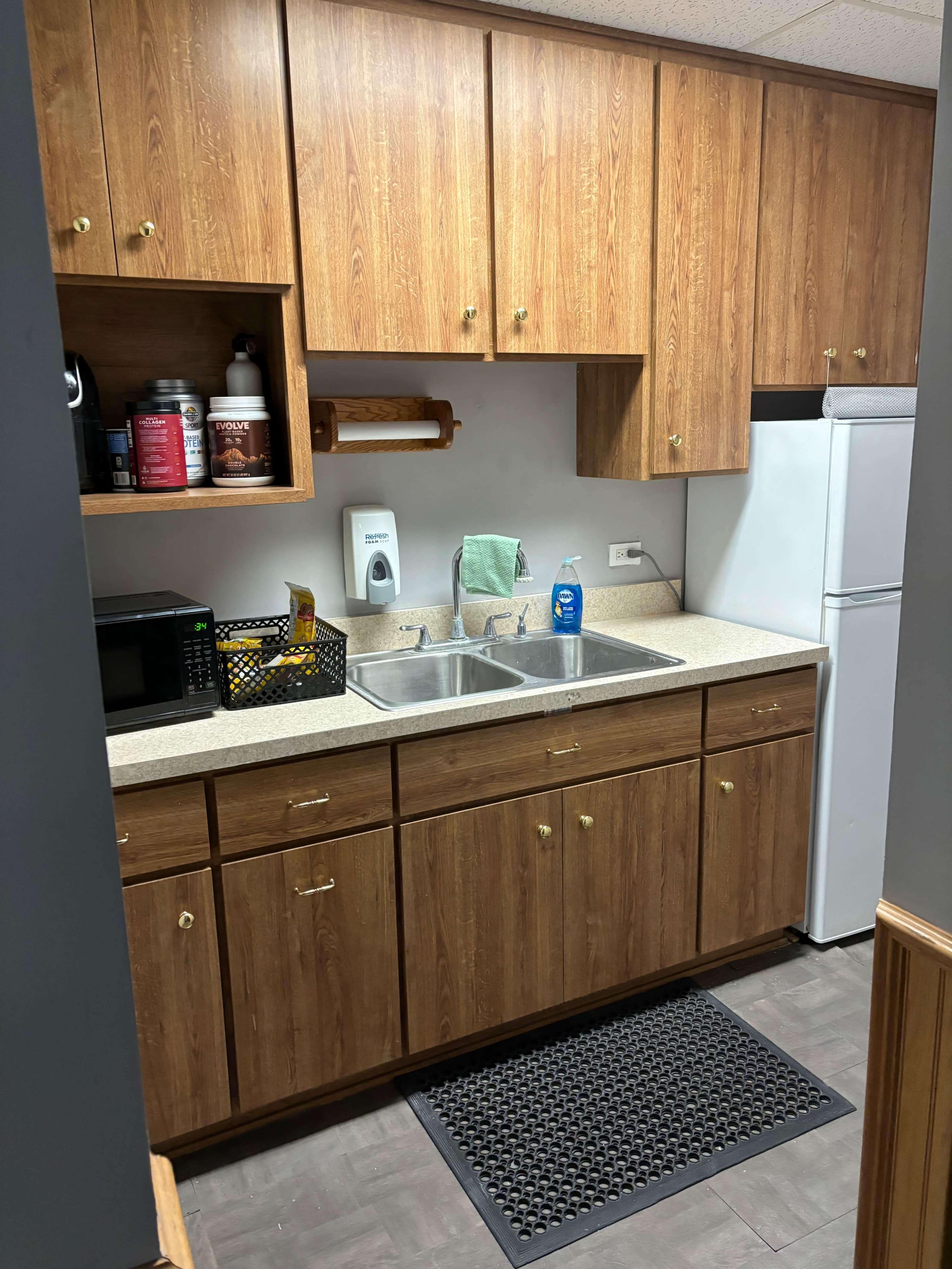 A kitchen area featuring wooden cabinets, a double sink, a countertop with various kitchen items, and a white refrigerator.