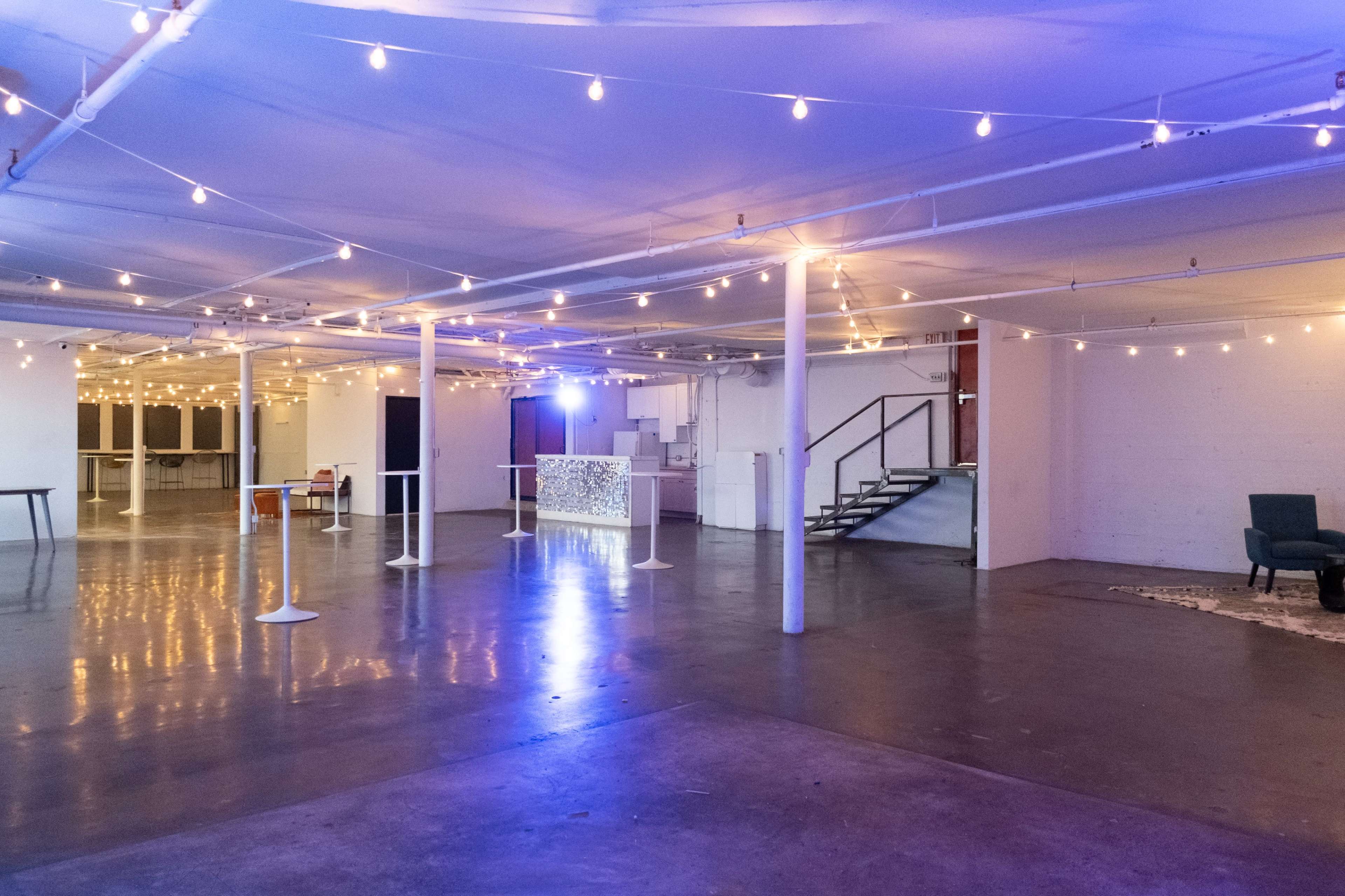 A spacious indoor area is illuminated by string lights hanging from the ceiling, with a staircase and various tables arranged throughout the space.