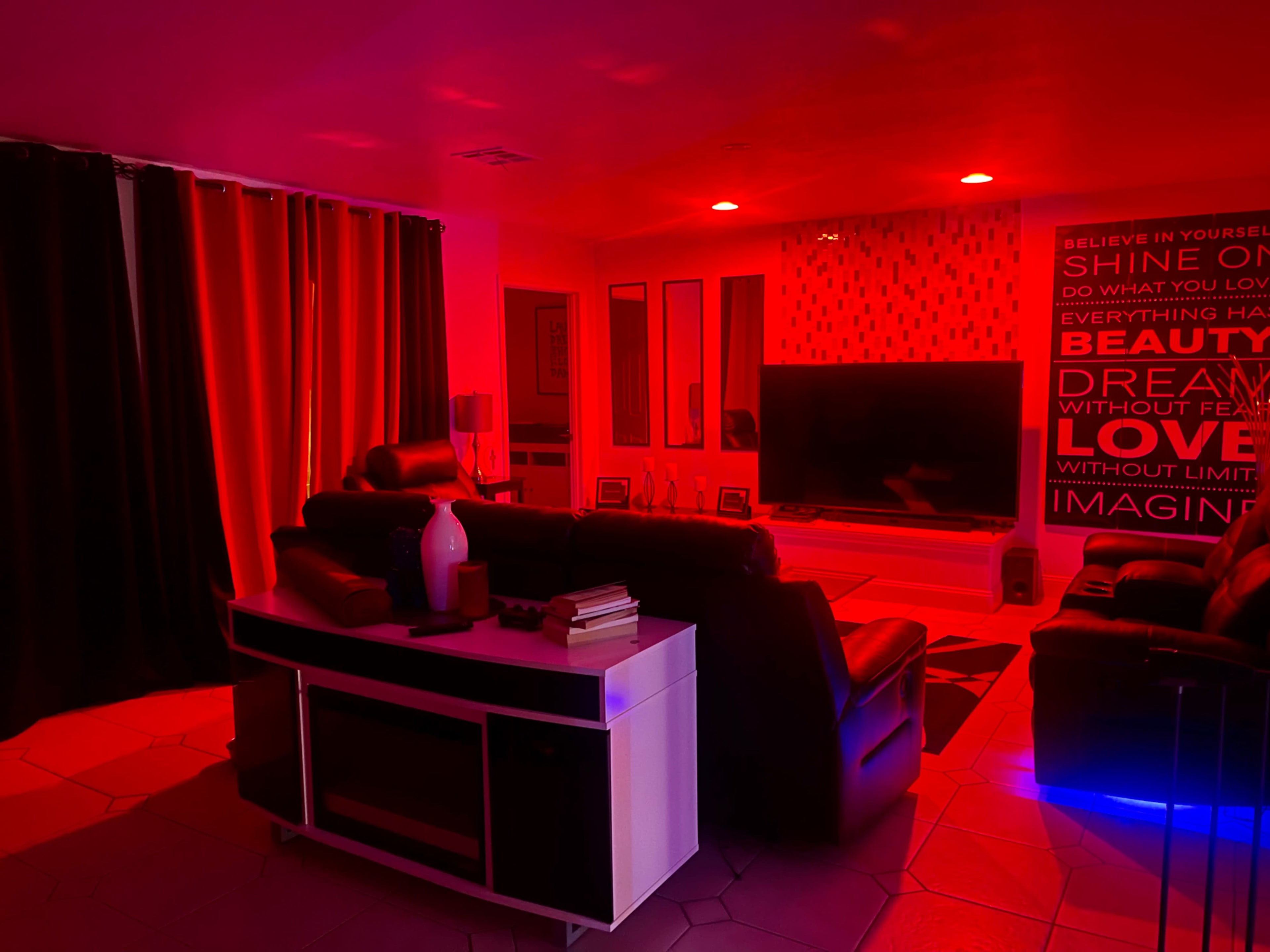 A living room features black recliners, a large television, and a wall adorned with motivational phrases, all illuminated by red lighting.