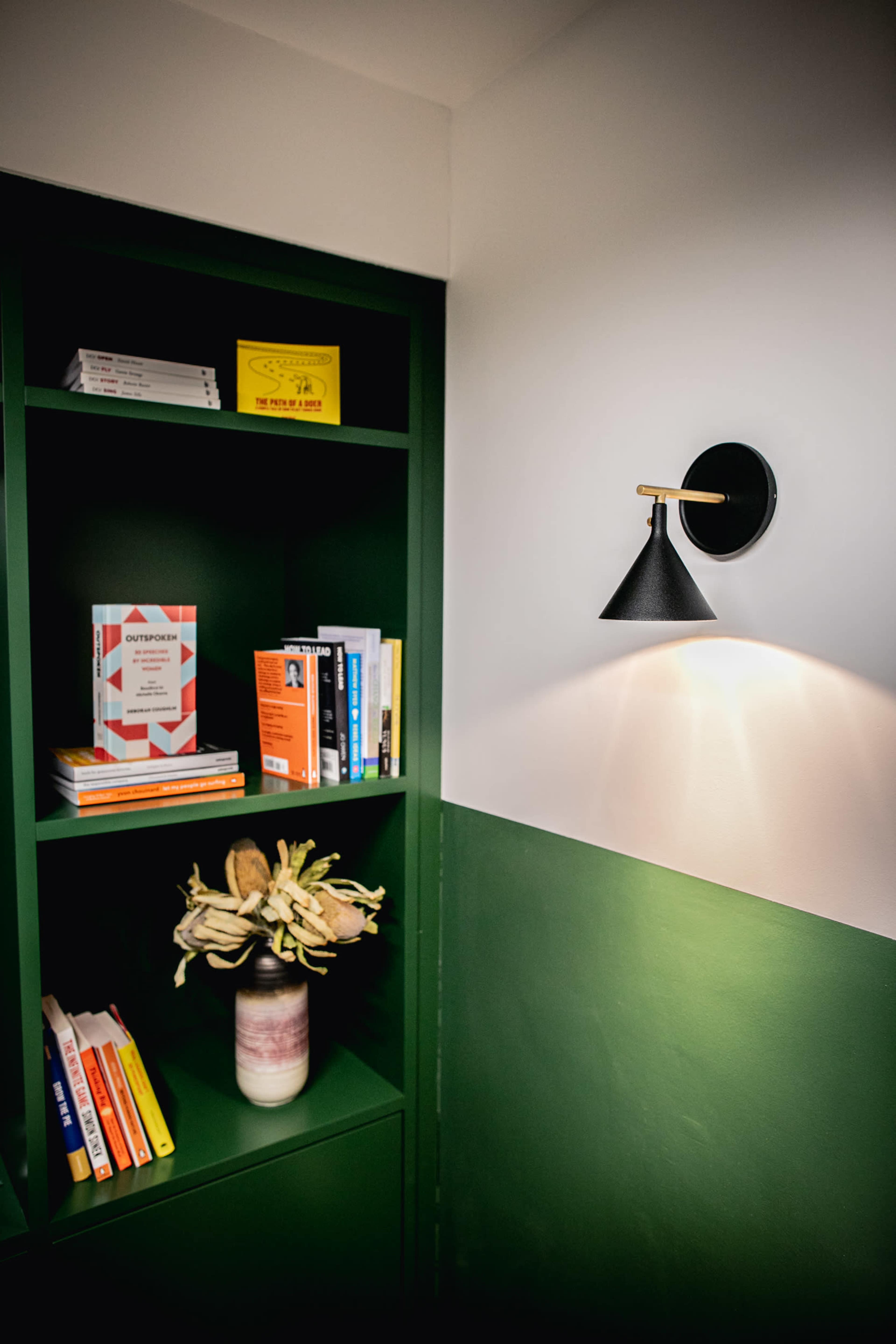 A corner of a room features a dark green bookshelf filled with books and decorative items, alongside a black wall sconce illuminating the space.