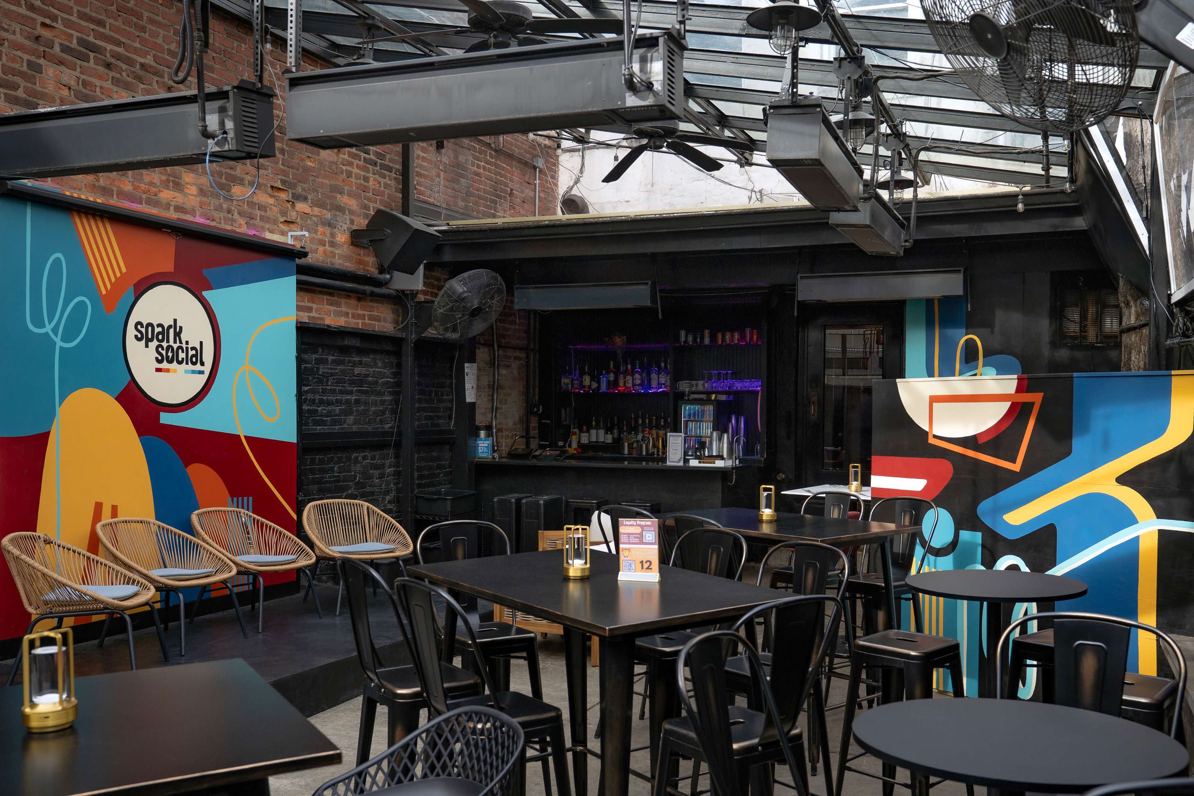 The image shows an outdoor seating area of a bar or restaurant with colorful murals, black tables, and rattan chairs under a glass roof.