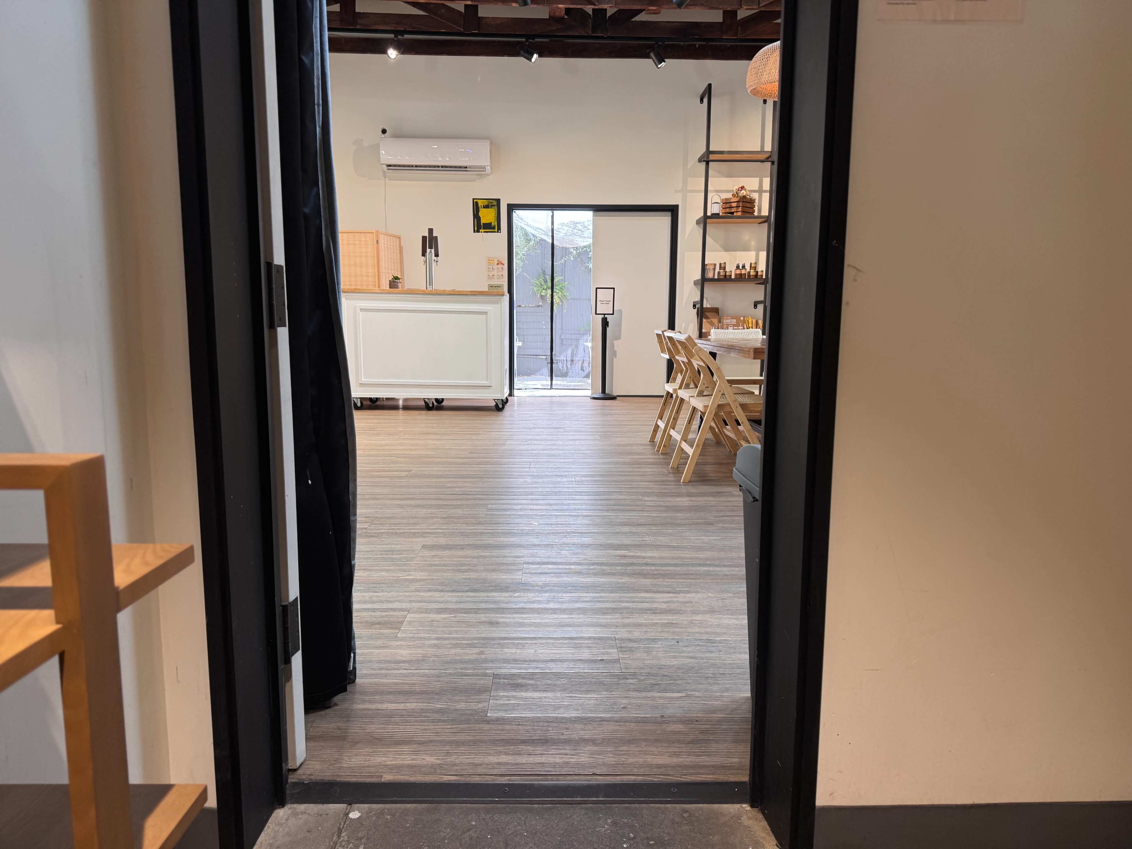 The image shows an open doorway leading into a light-colored interior space with wooden flooring, showcasing tables and chairs arranged neatly.