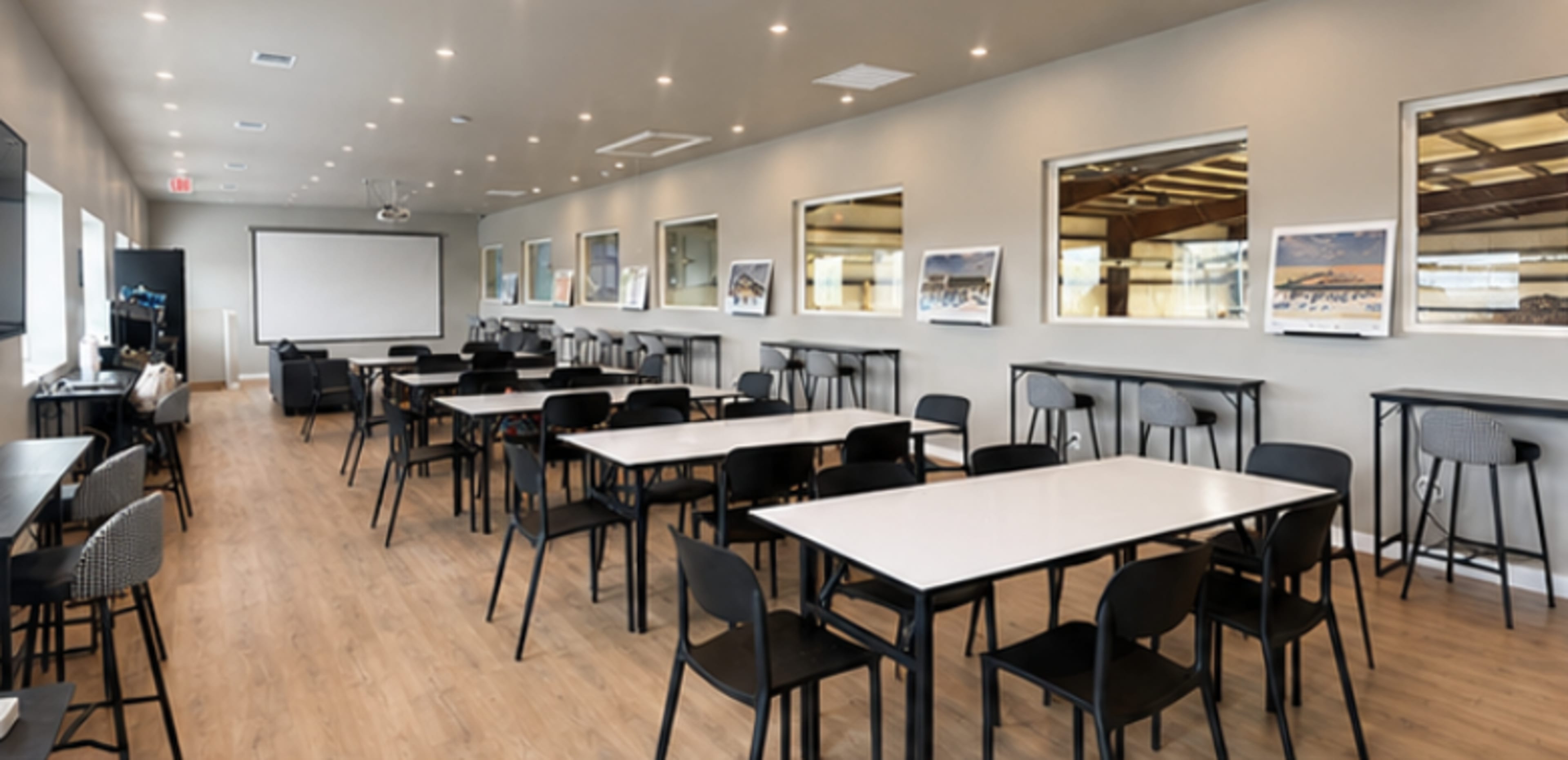 The image shows a spacious meeting room with multiple rectangular tables and black chairs arranged neatly, featuring large windows and a projector screen.