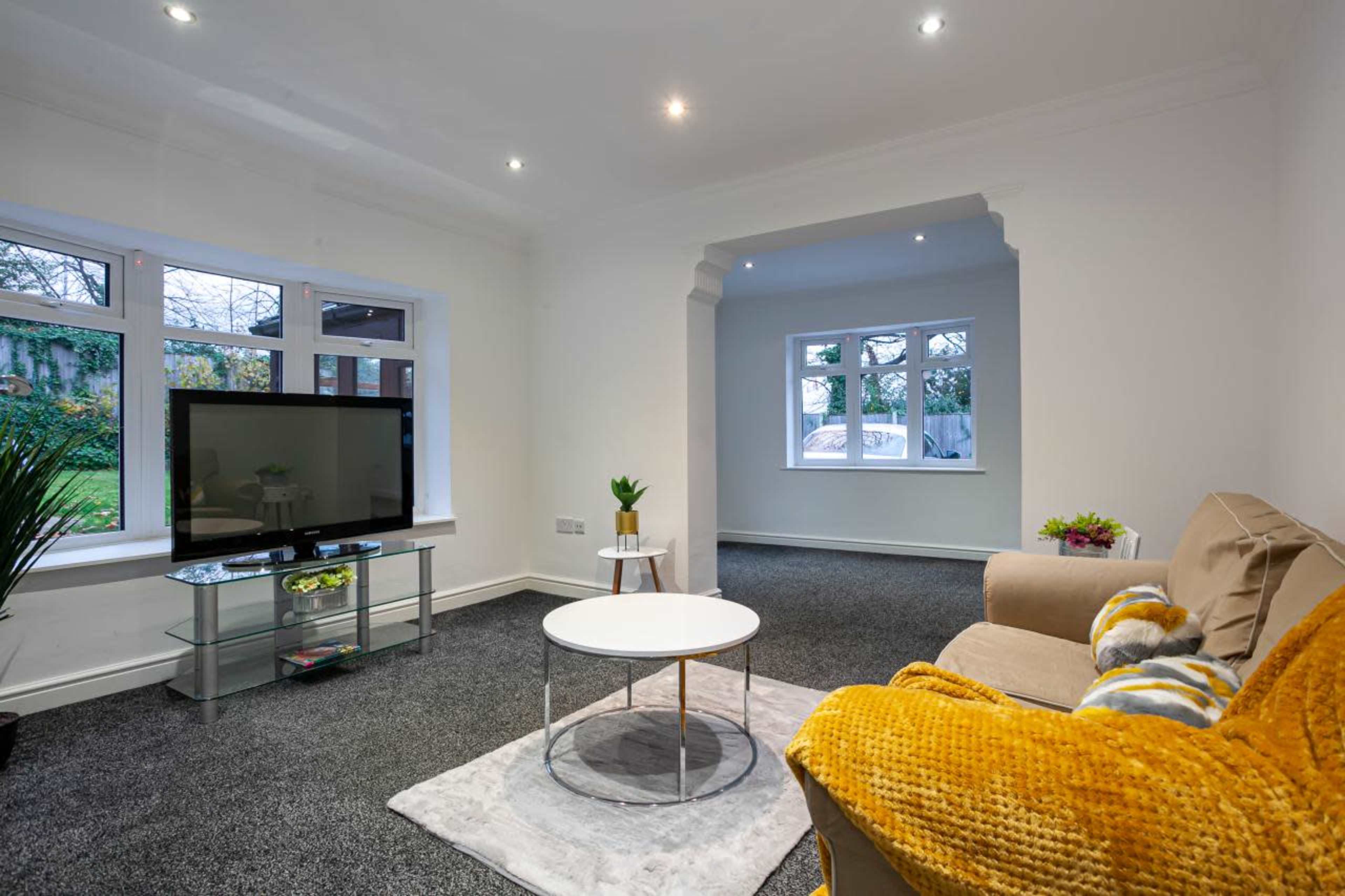 A well-lit living room featuring a beige sofa, a round coffee table, a television on a glass stand, and large windows allowing natural light to enter.