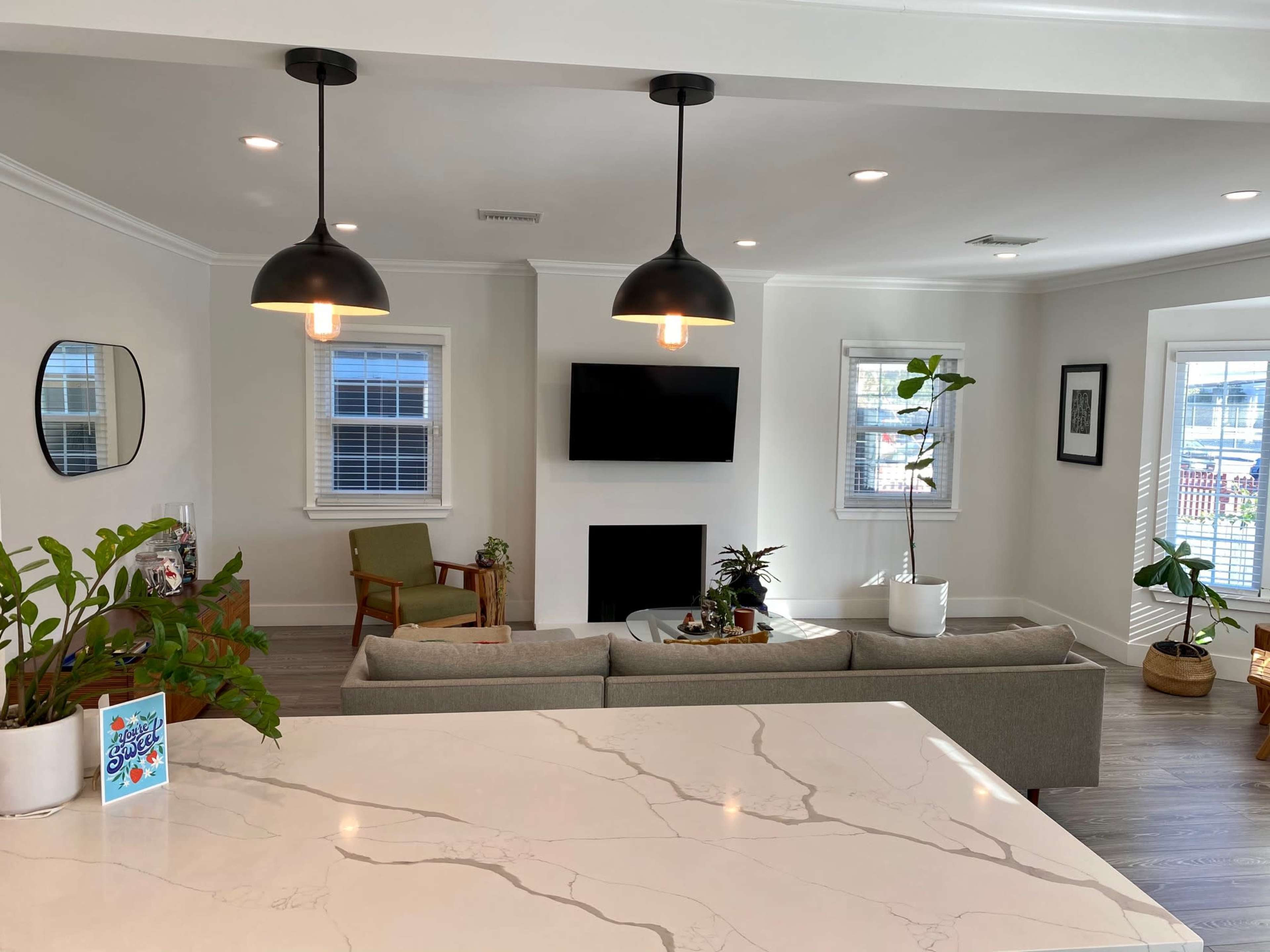 A modern living room features a sofa, green plants, a television mounted on the wall, and pendant lights hanging from the ceiling.
