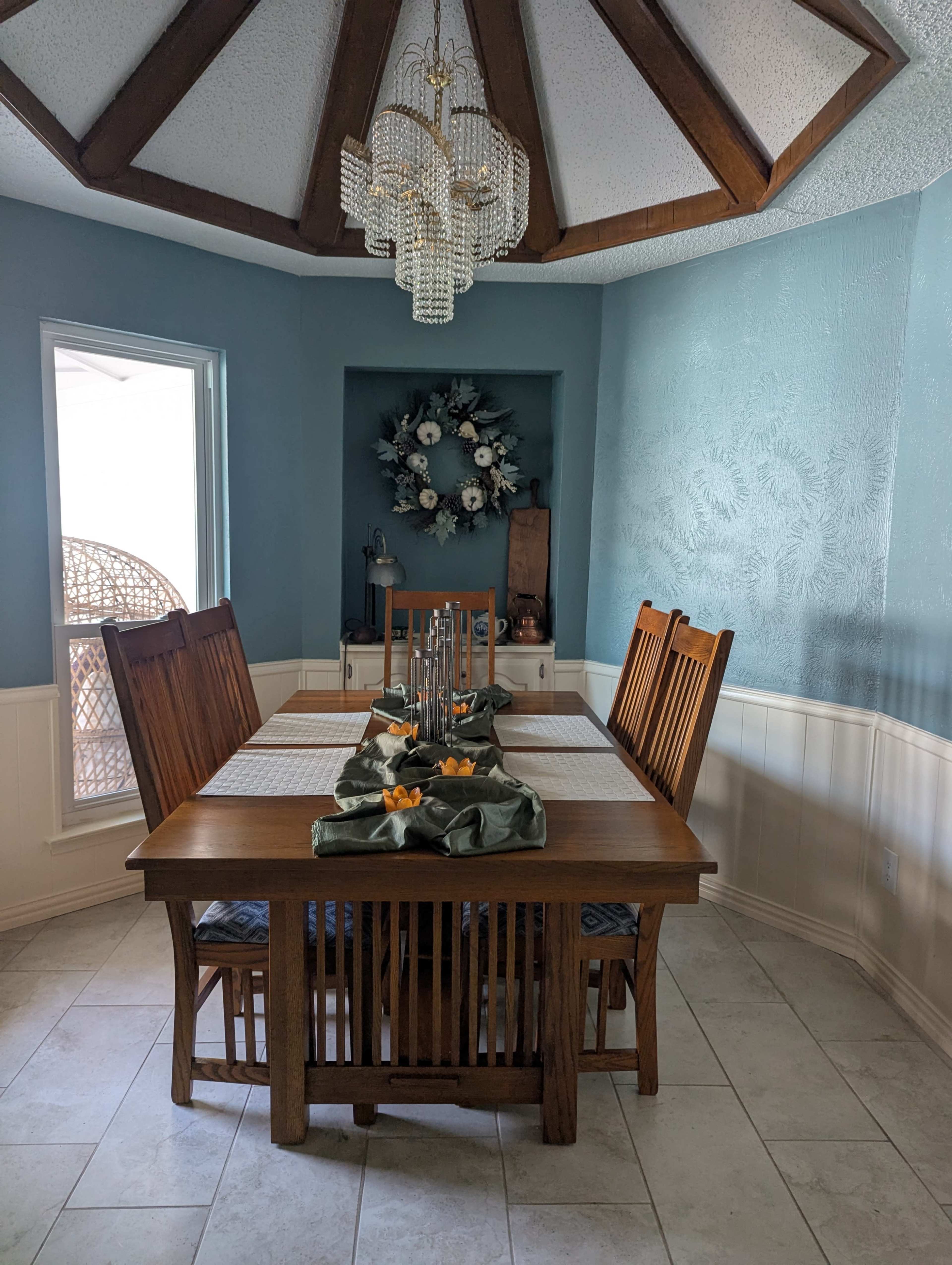 A dining room features a wooden table set for six with a centerpiece and a chandelier above, surrounded by blue walls and a large window.